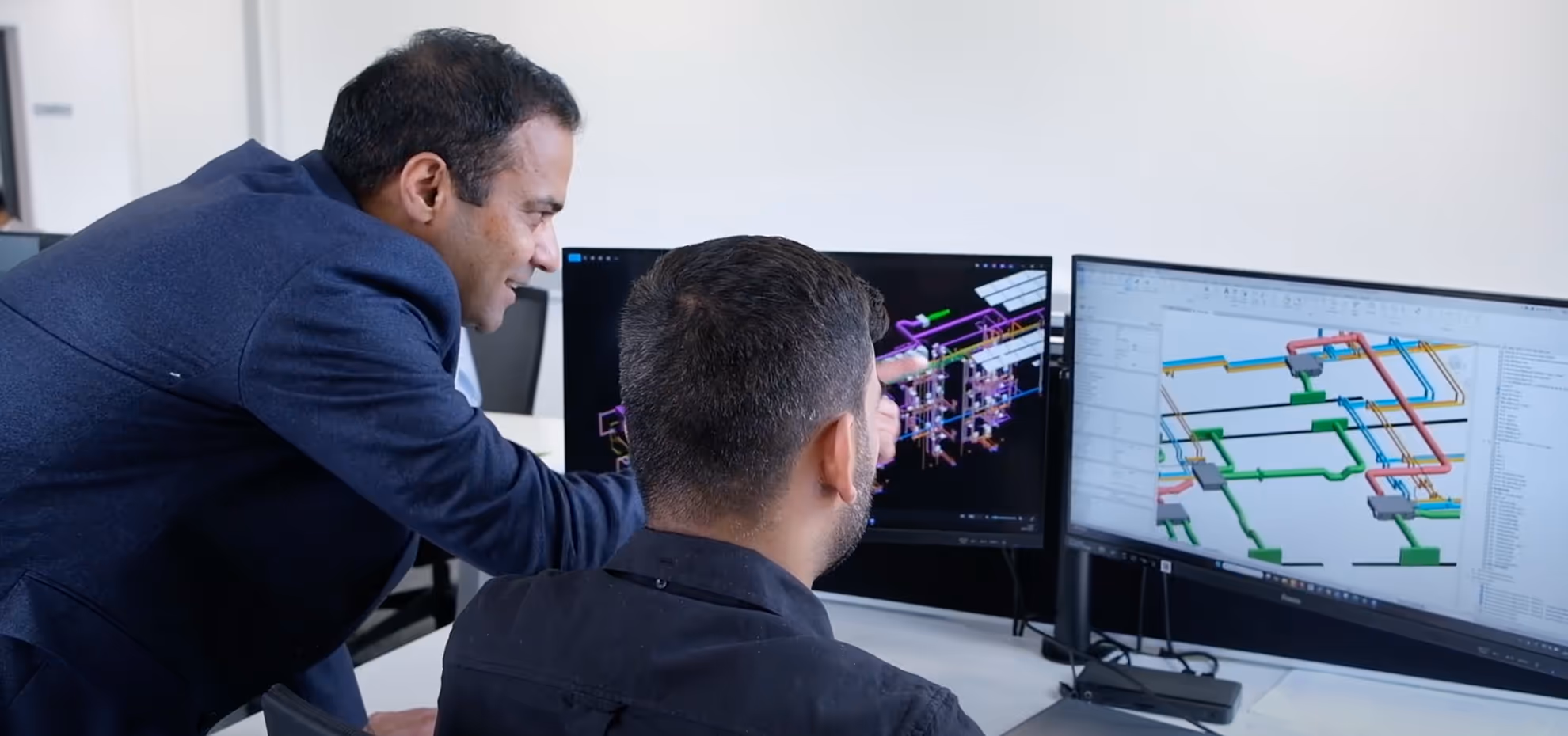 Two men collaborating over complex 3D engineering models displayed on dual computer monitors in an office.
