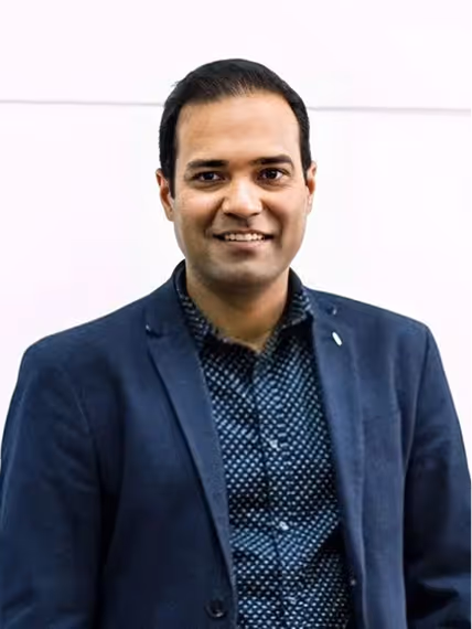 Smiling man wearing a dark blue blazer and patterned shirt against a plain light background.