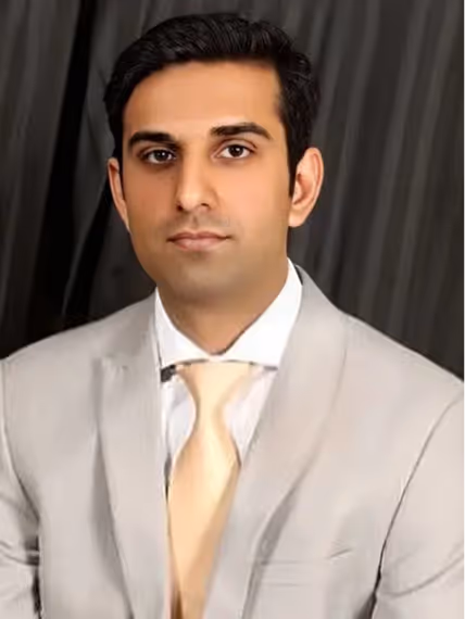 Portrait of a man wearing a light gray suit jacket, white shirt, and pale yellow tie against a dark curtain background.