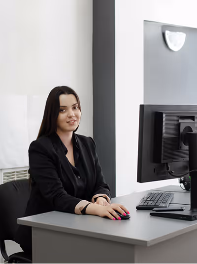 Smiling woman with long dark hair sitting at a desk using a computer mouse, facing slightly toward the camera.