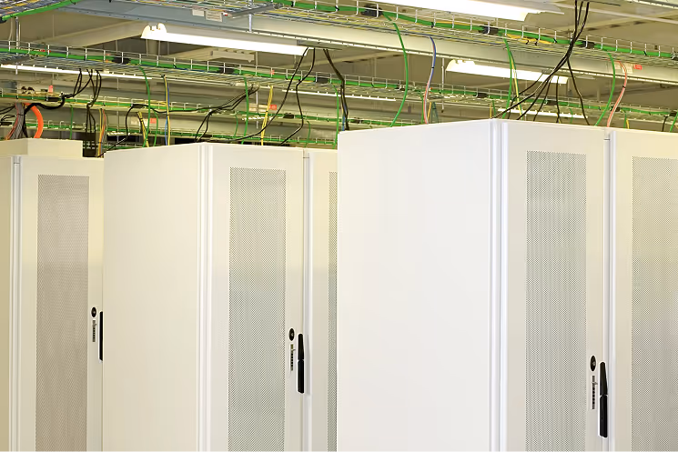 Rows of white server racks in a data center with exposed ceiling cables and fluorescent lighting.