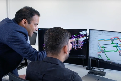 Two men reviewing complex 3D engineering designs on dual computer monitors in an office setting.