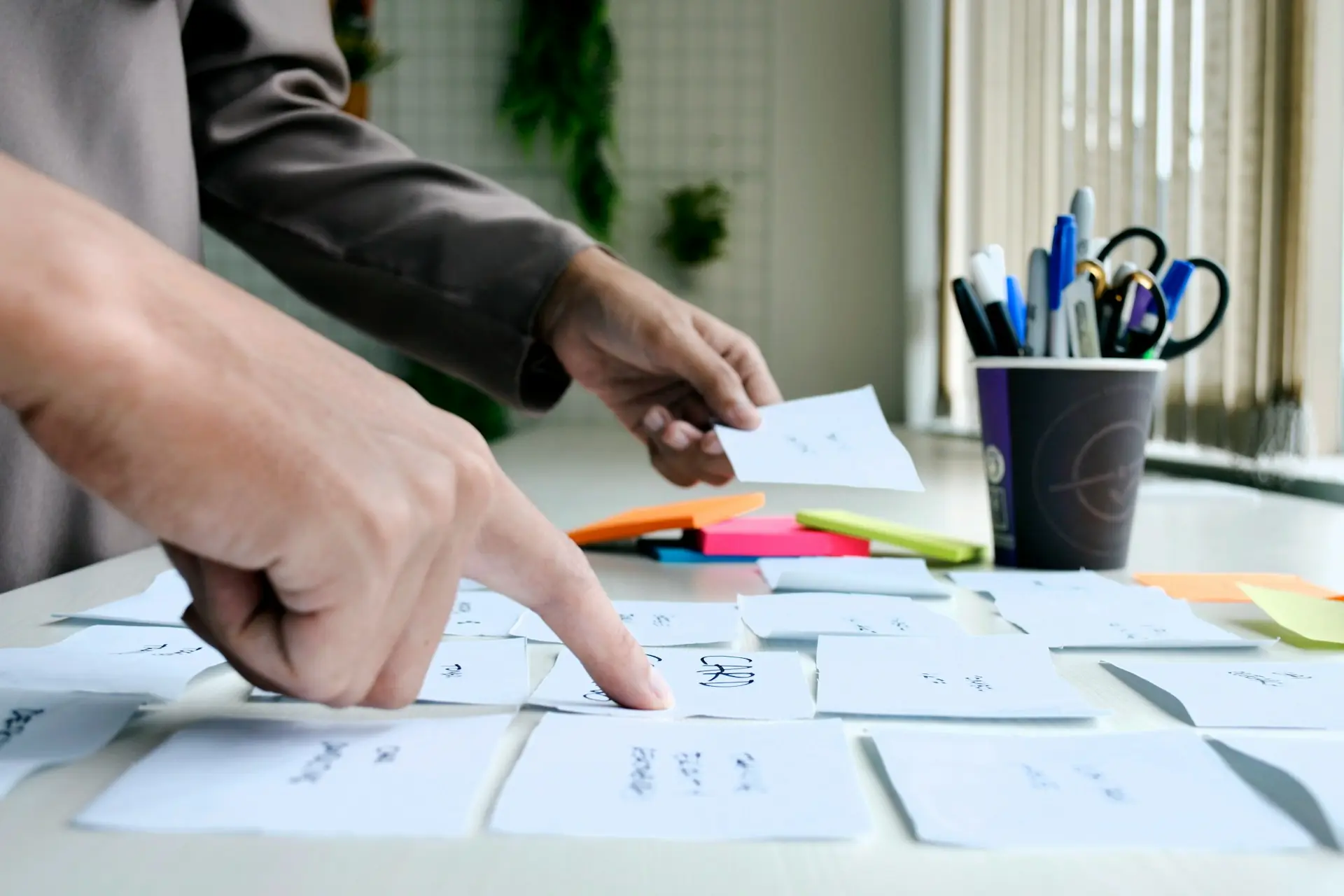 Person organizing white paper notes on a table with colorful sticky notes and a cup holding pens and scissors.