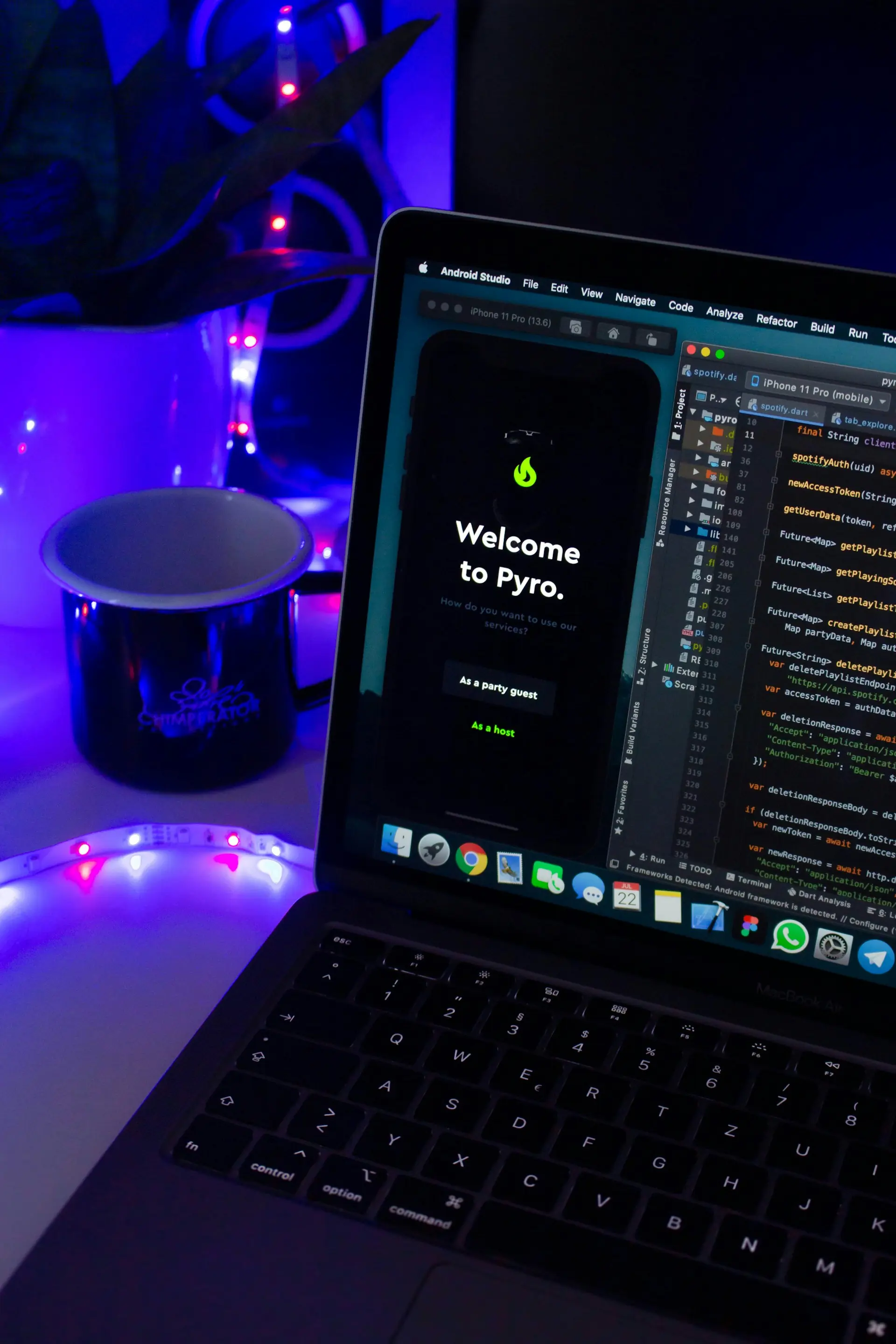 Laptop displaying Android Studio with code and a mobile app welcome screen saying 'Welcome to Pyro' in a dim room with blue and purple ambient lighting, a plant, and a mug nearby.