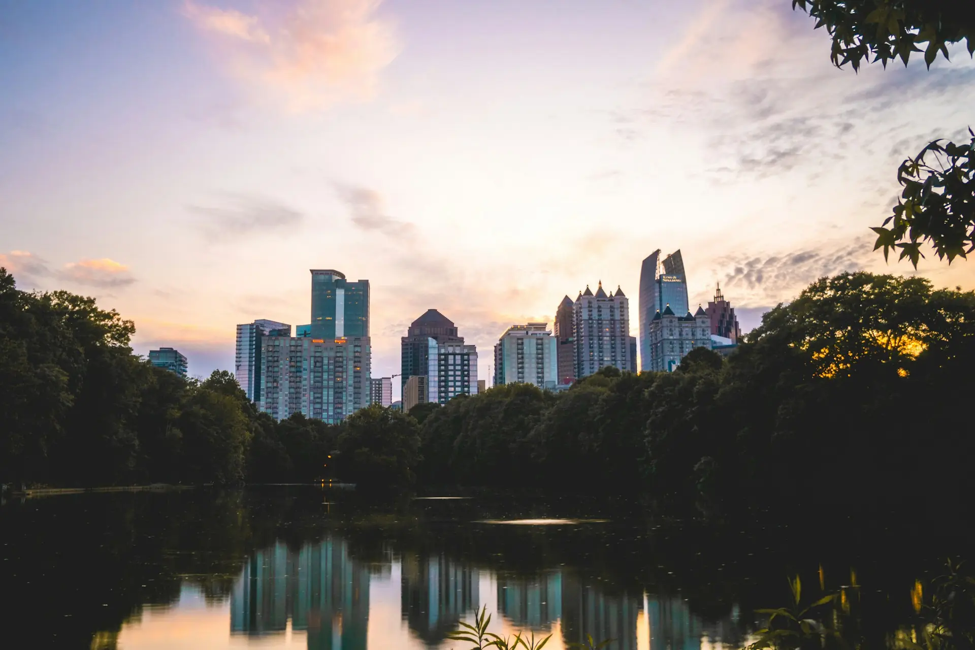 City skyline with modern high-rise buildings reflected in a calm river at sunset, framed by trees.