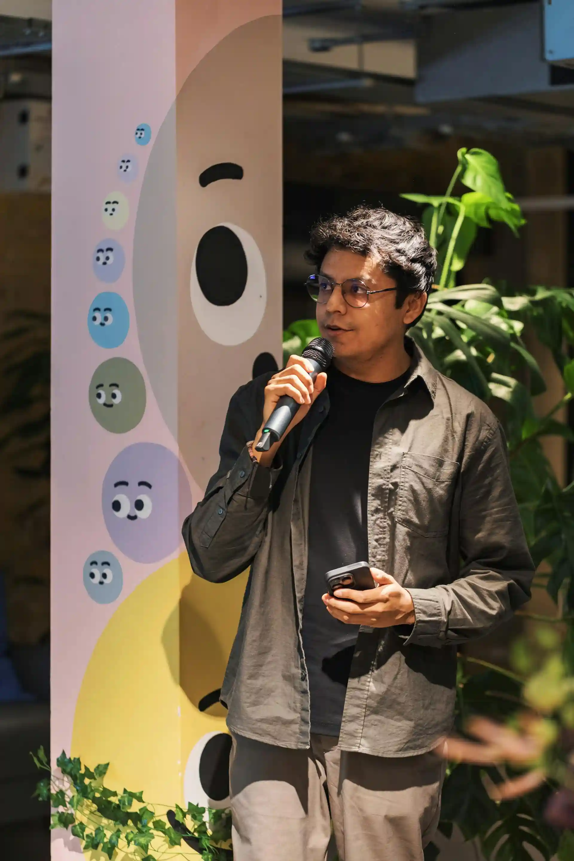 A man wearing glasses and a dark shirt speaking into a microphone while holding a smartphone, standing next to a colorful column with painted cartoon faces and surrounded by green plants.