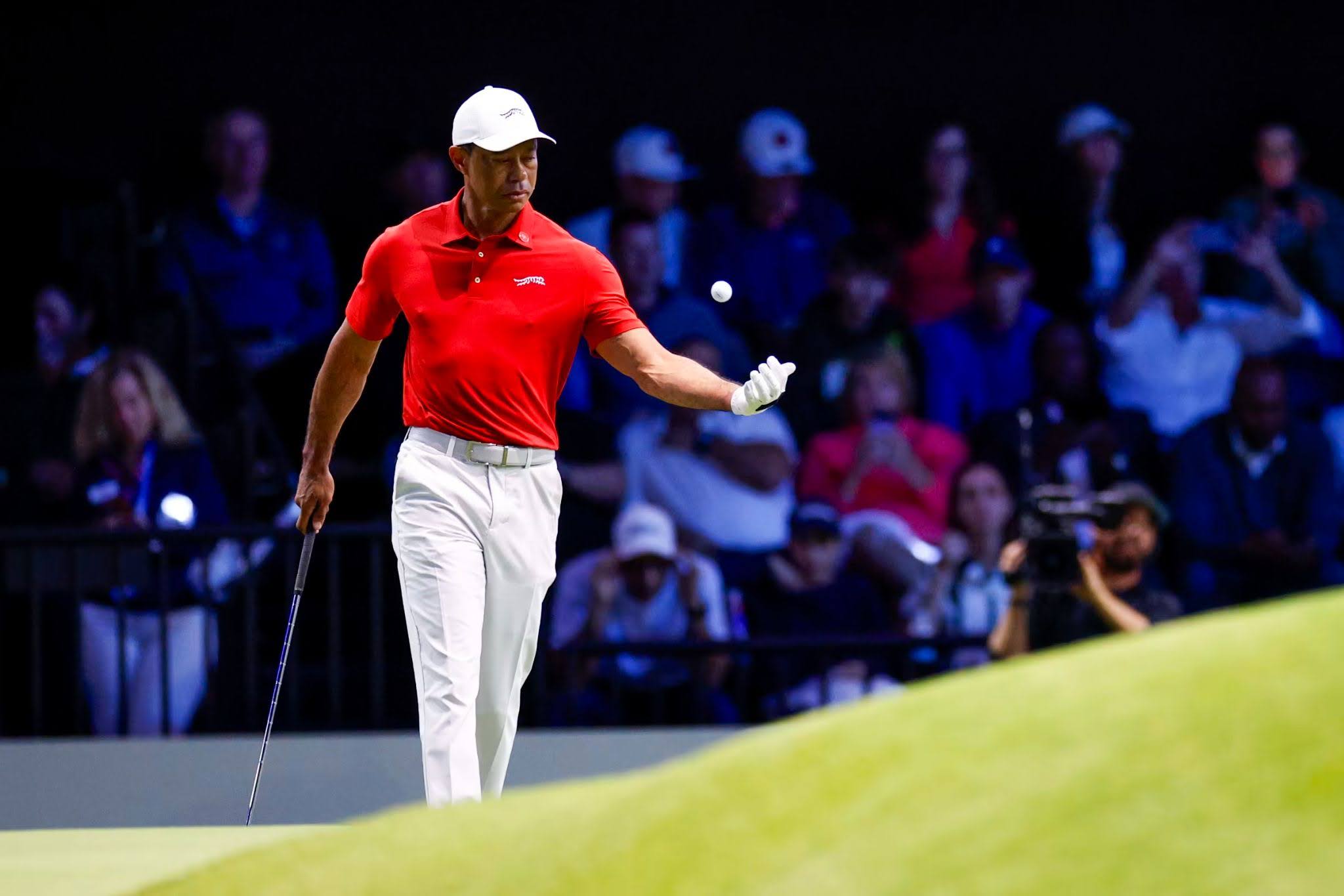 Golfer in a red shirt and white pants tossing a golf ball on the green with spectators watching in the background.