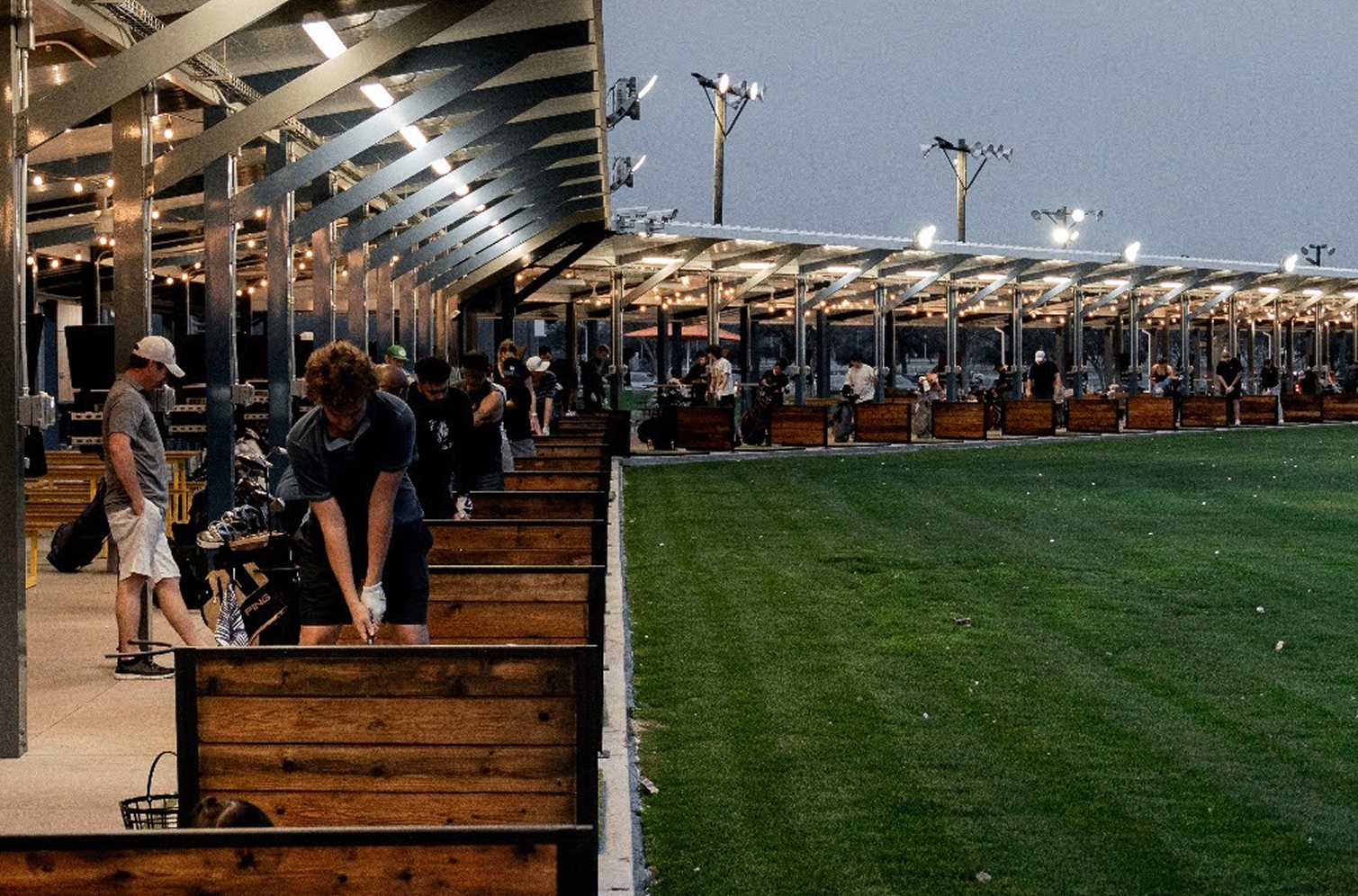 People practicing golf swings at an outdoor driving range with individual wooden stalls and overhead lights at dusk.