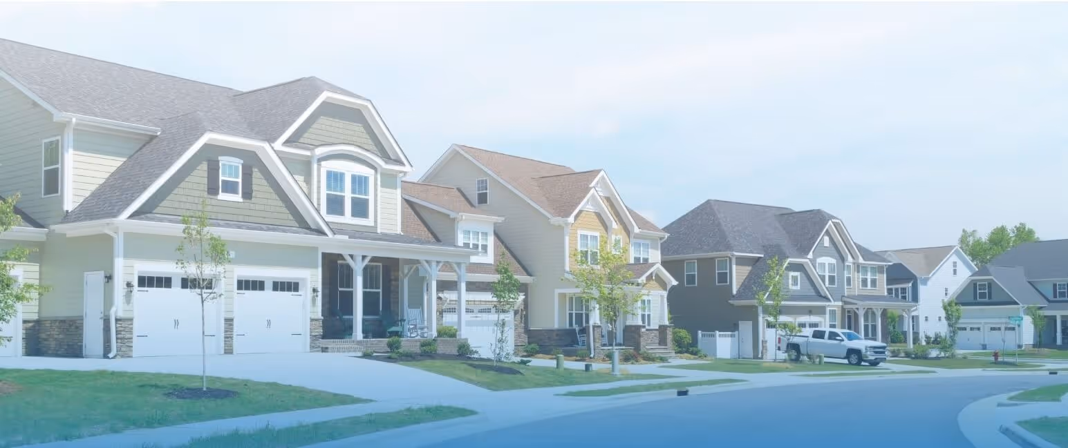 Suburban street view with several large two-story houses featuring garages, front porches, and well-kept lawns.