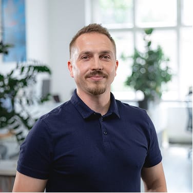 Muž s krátkými vlasy a knírkem v tmavě modrém polo tričku stojí v moderní světlé kanceláři s rostlinami v pozadí.