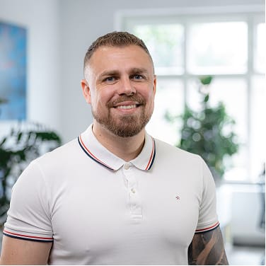 Usměvavý muž s krátkými vlasy a vousy v bílé polokošili stojí v interiéru s rozostřeným světlým pozadím.