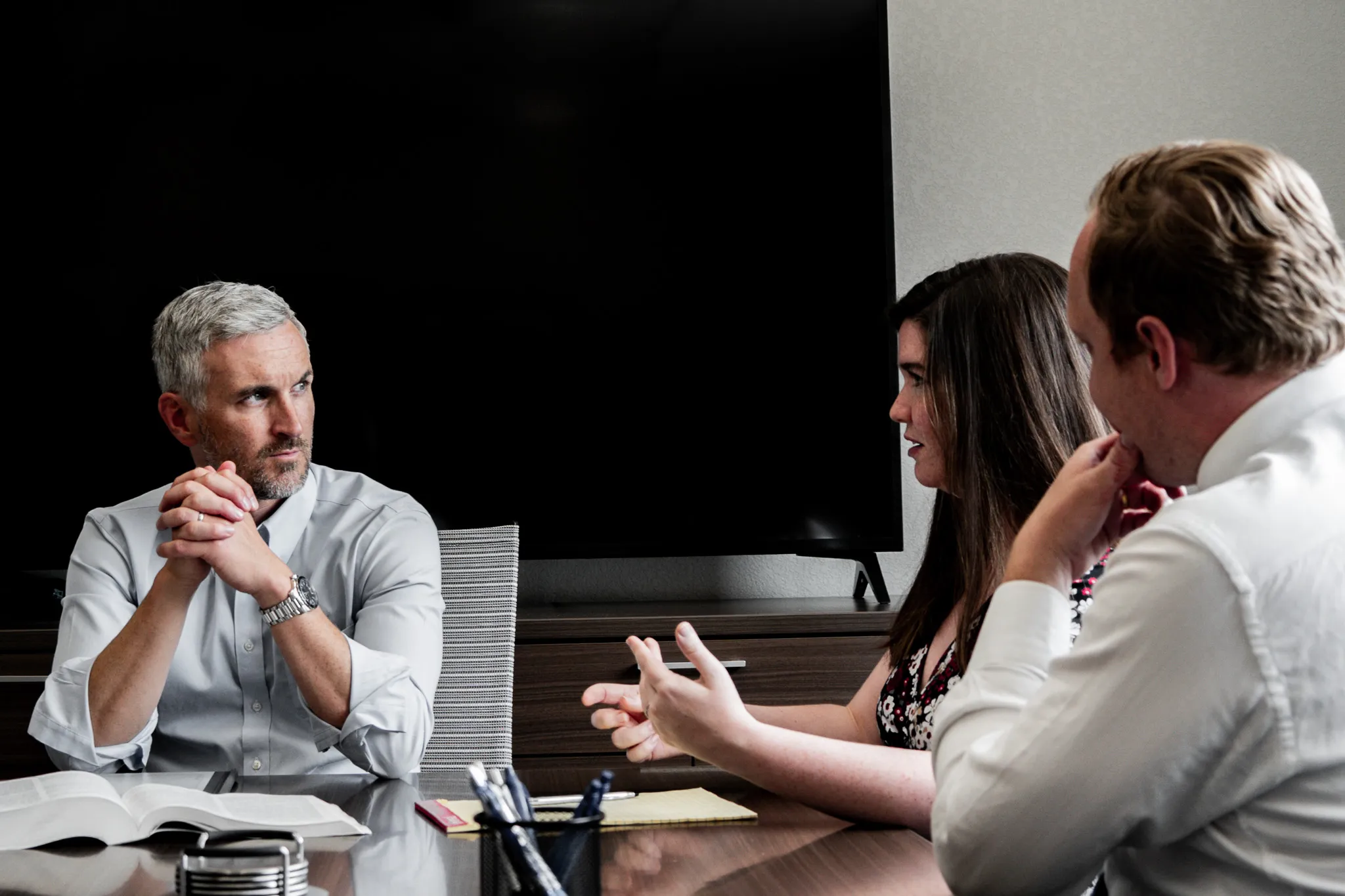 Three businesspeople in a meeting discussing around a table with an open book and notepad.