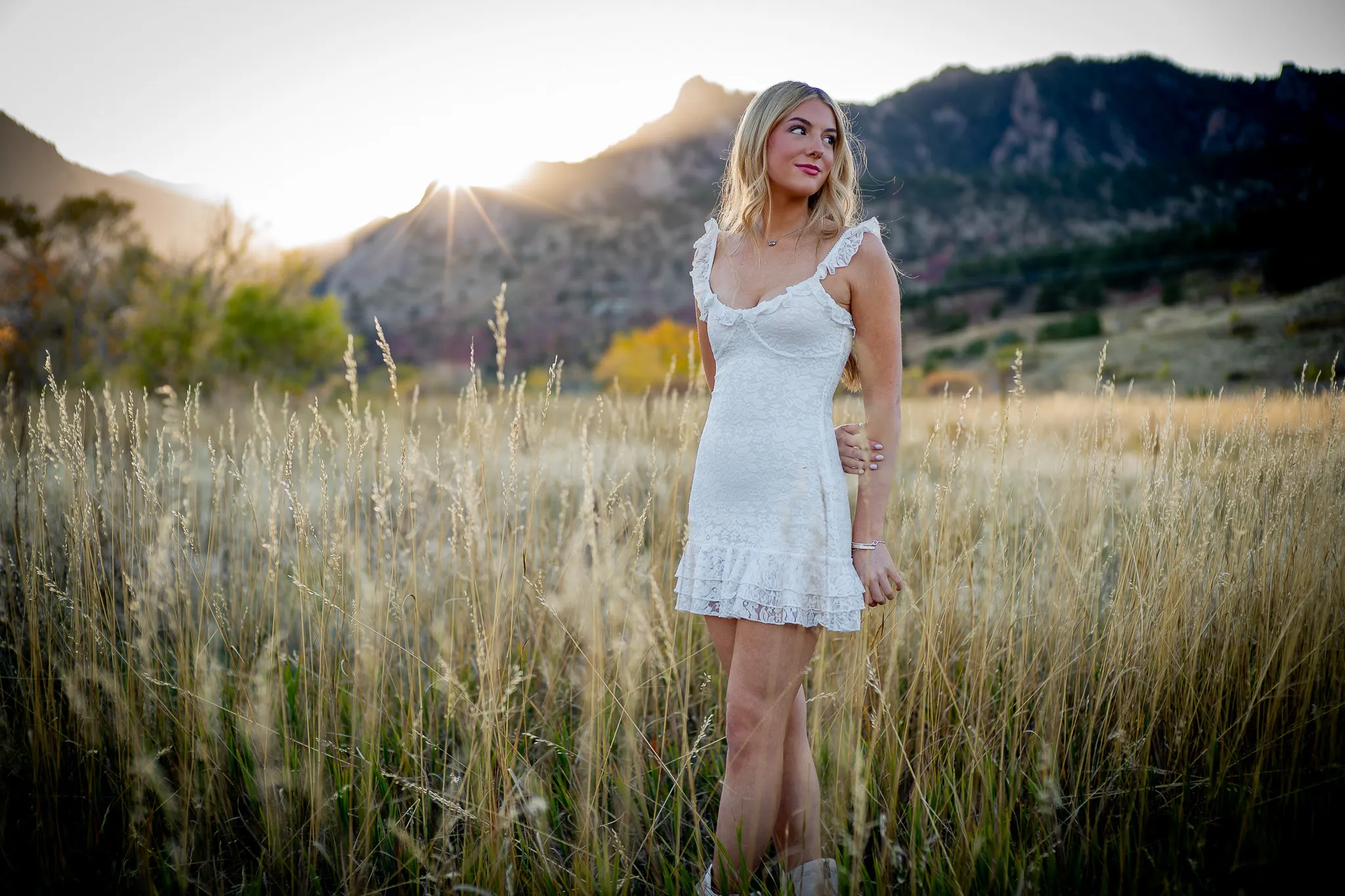 Young woman in a white dress standing in a grassy field with mountains and sunset in the background.