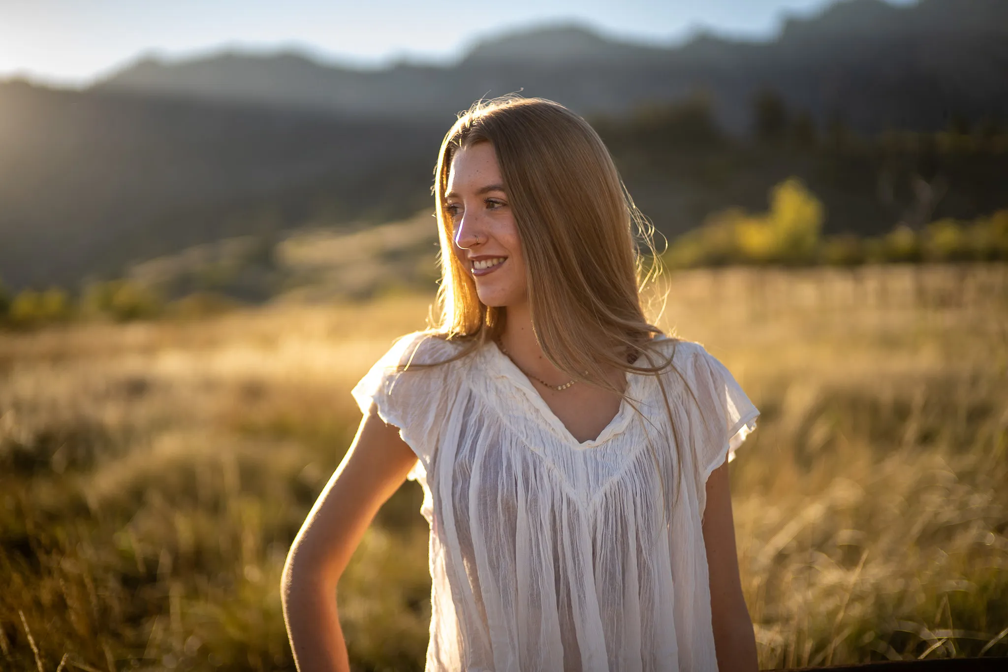 Young woman with long hair wearing a white blouse smiling and looking to the side in a sunlit grassy field with mountains in the background.