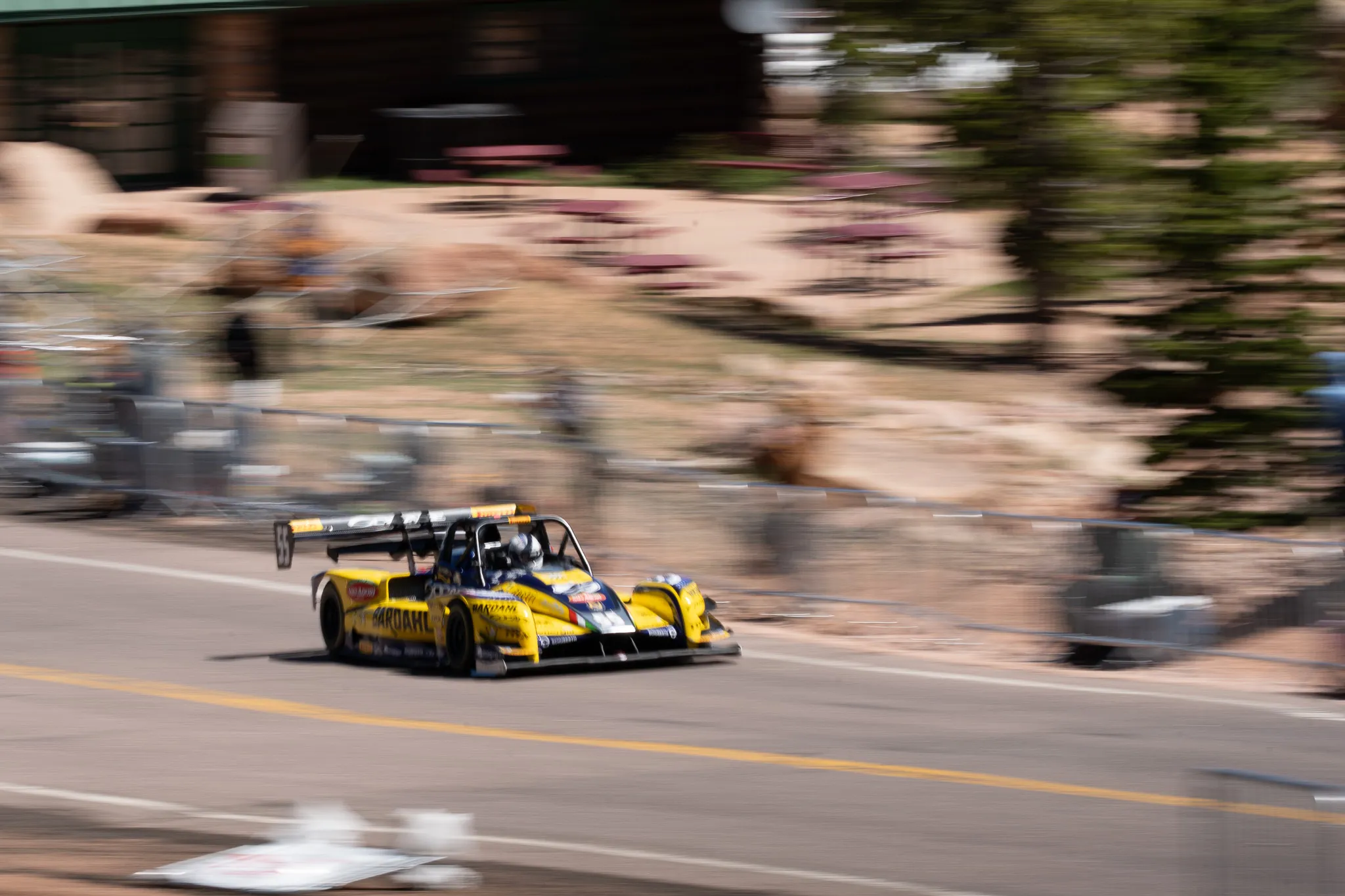 Yellow race car with spoiler speeding on a paved road with blurred background of trees and barriers taken by Cody Strate