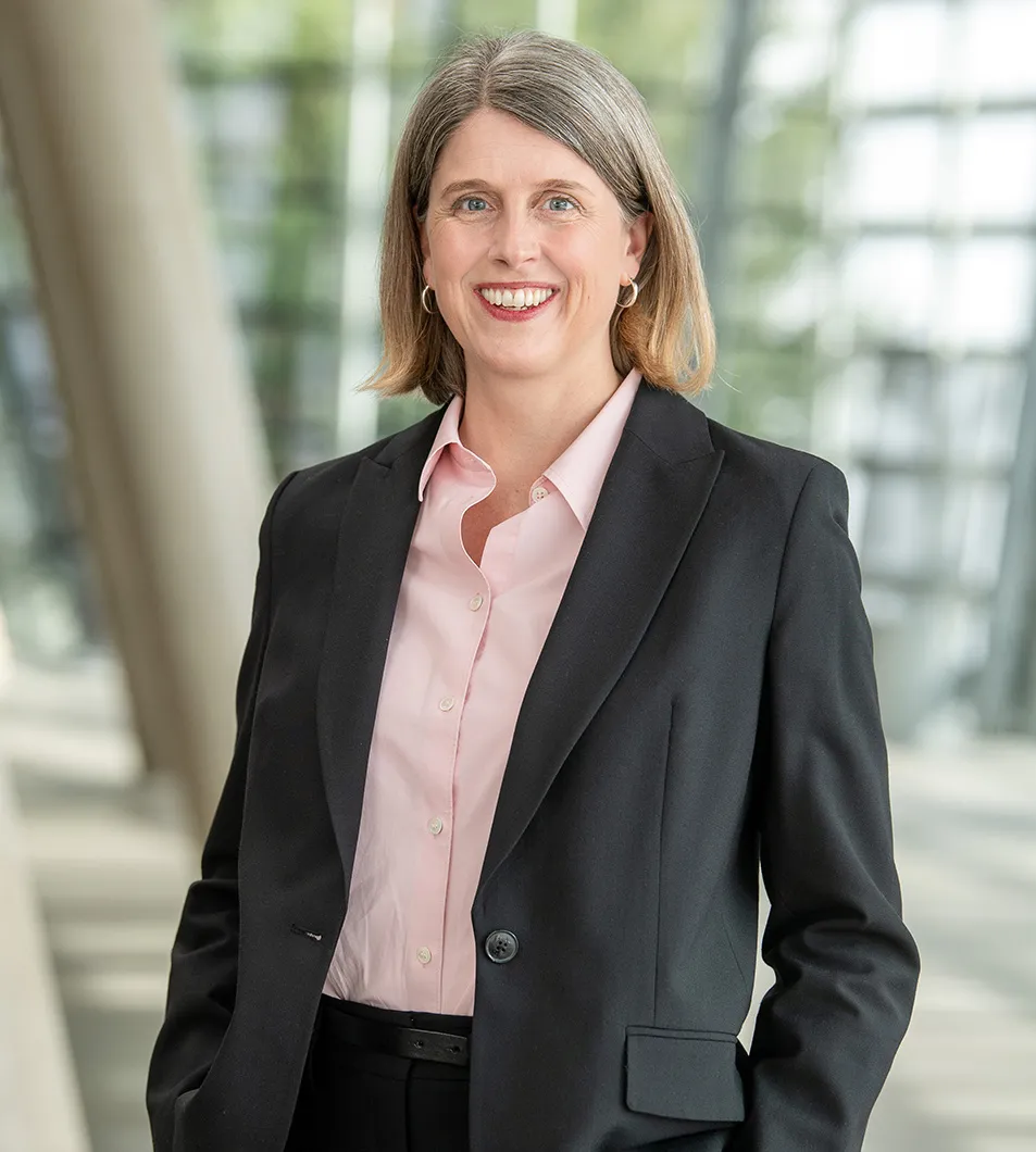 Smiling woman with shoulder-length gray hair wearing a black blazer and light pink button-up shirt in a modern indoor setting.