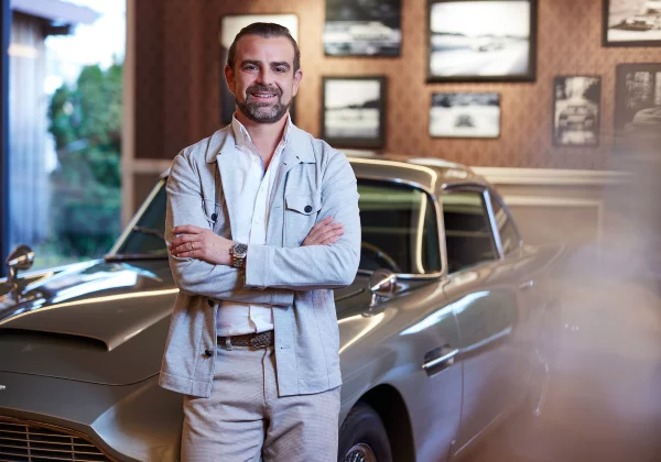 Smiling man with arms crossed standing in front of a vintage car in a warmly lit room with framed pictures on the wall.