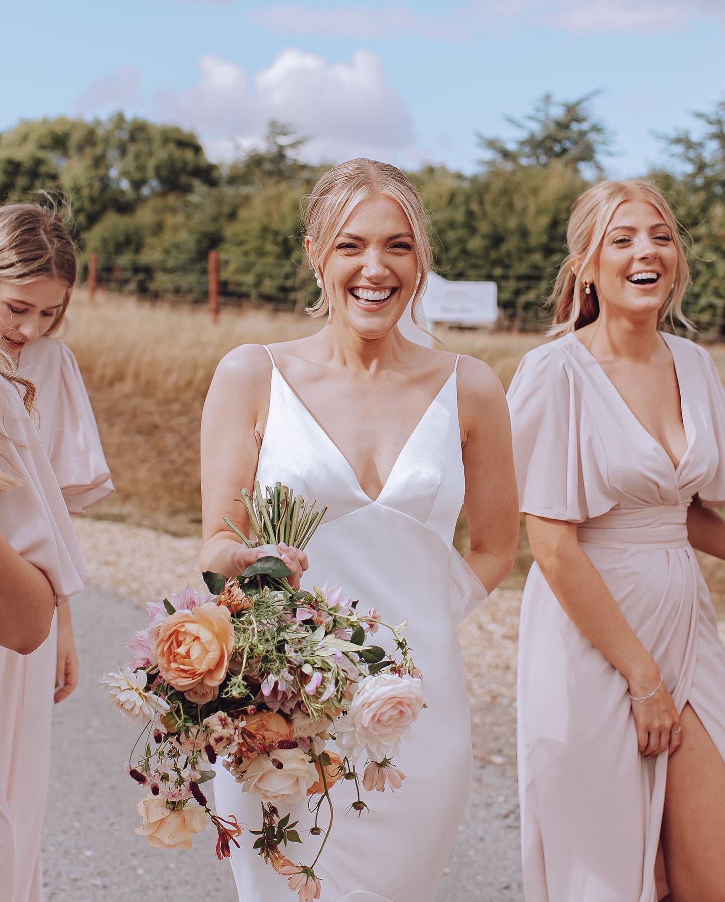 Smiling bride in a white dress holding a large bouquet of flowers, standing outdoors with bridesmaids in light pink dresses.