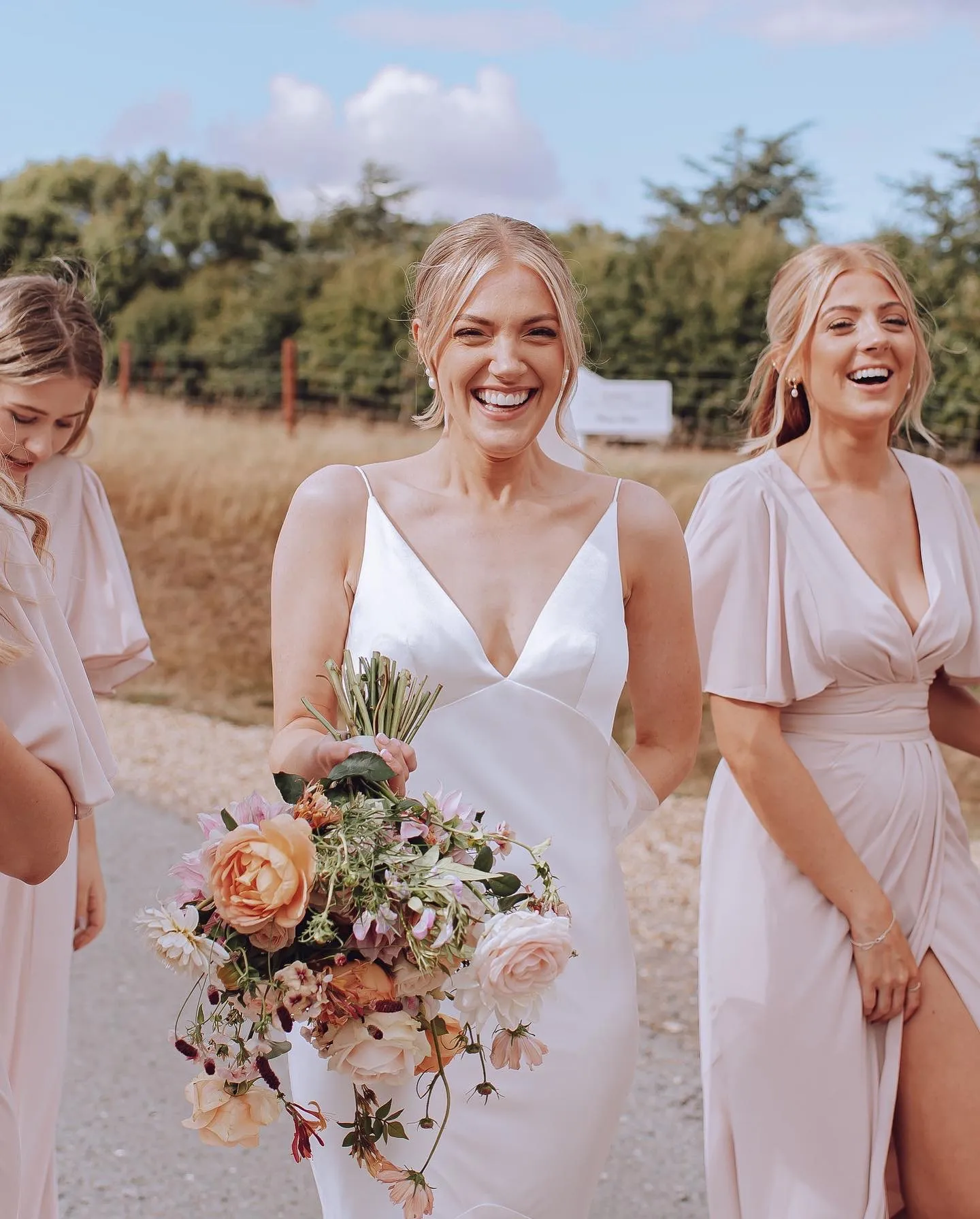 Bride in white dress holding a bouquet of mixed flowers, smiling with two bridesmaids in pale pink dresses outdoors.
