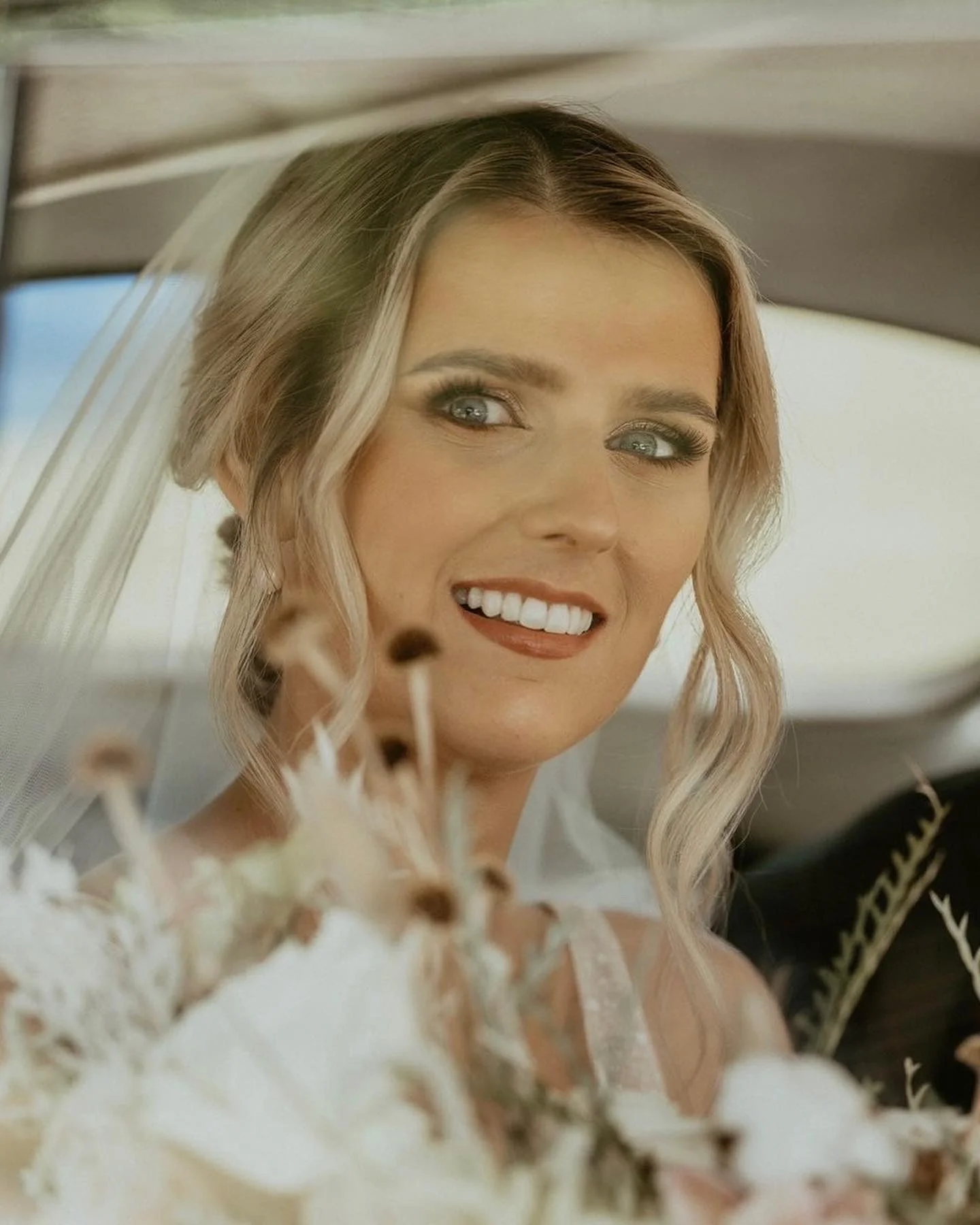 Smiling bride with wavy blonde hair and veil sitting in a car holding a bouquet of flowers.
