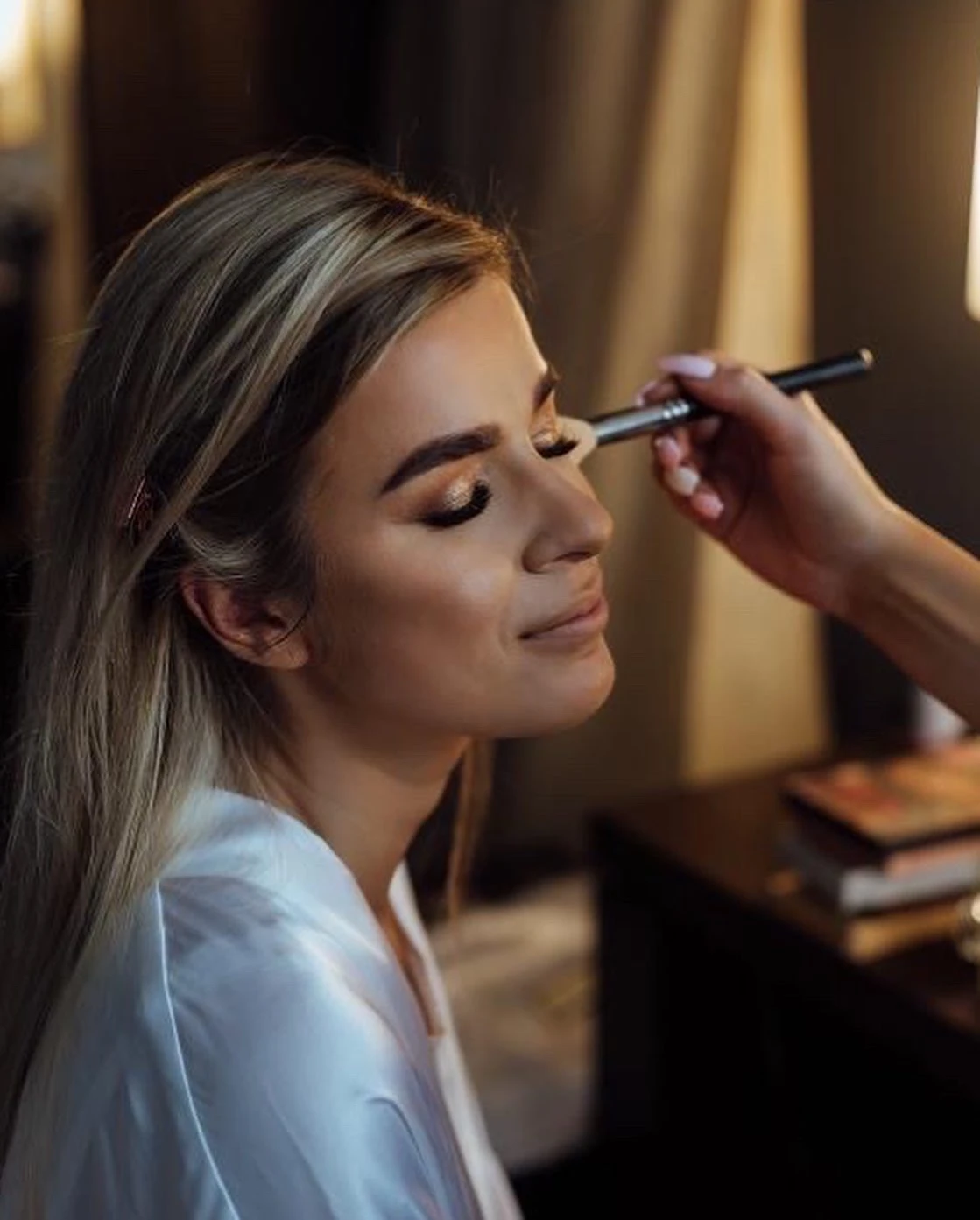Woman with closed eyes having makeup applied with a brush on her eyelids, wearing a white robe.