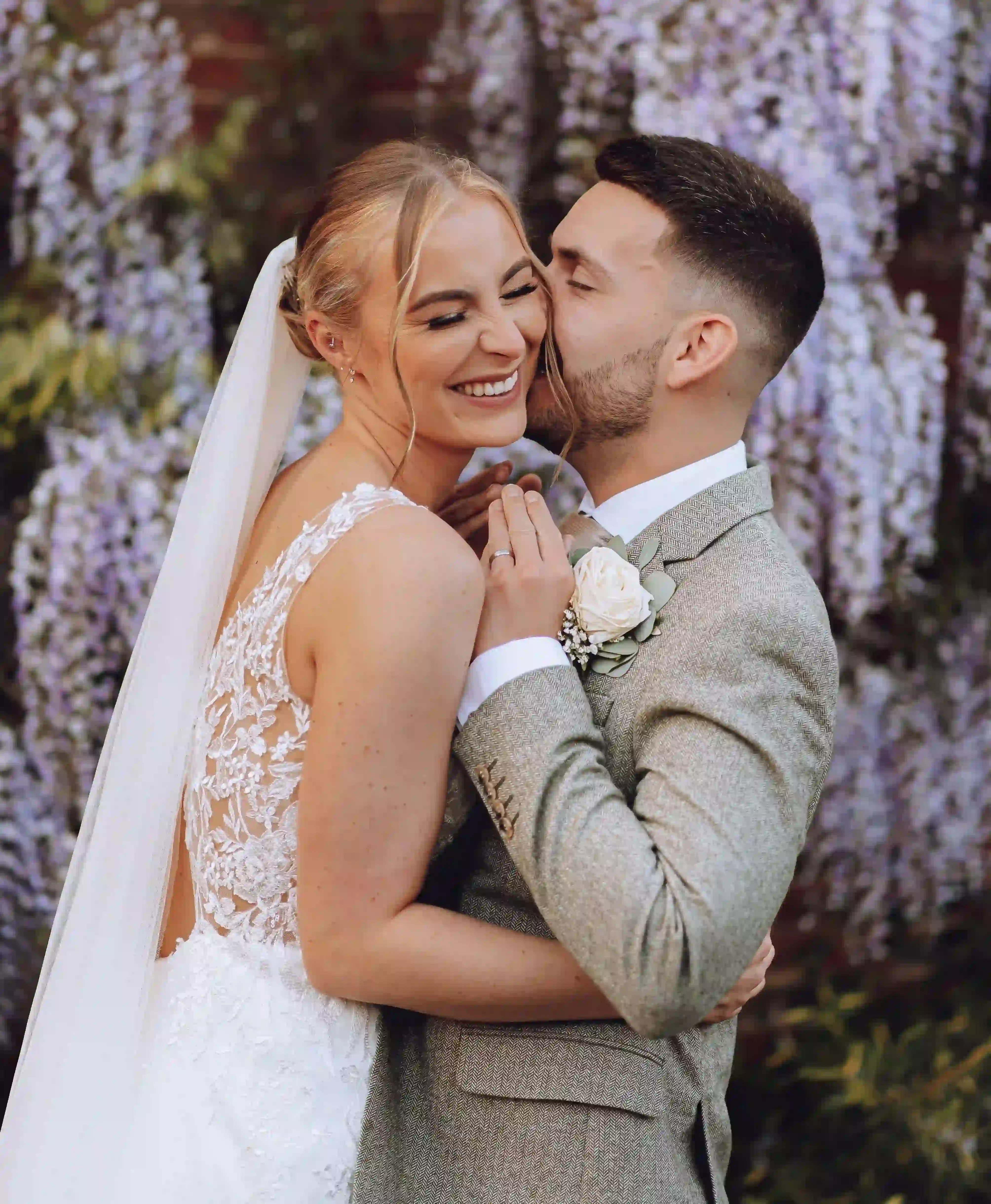 Bride smiling with eyes closed as groom kisses her cheek against a backdrop of purple flowers.