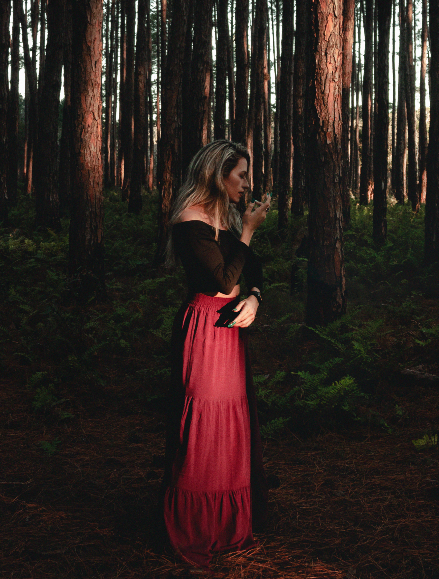 Woman in a black off-shoulder top and long red skirt standing contemplatively in a dense forest with tall trees.