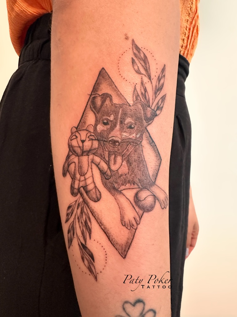 Tattoo of a happy dog holding a stuffed toy and a ball, with leaves and geometric shapes.