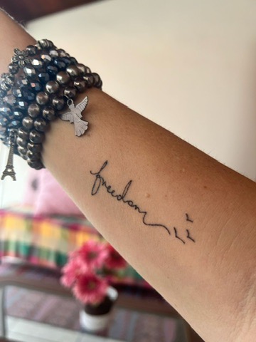 Forearm tattoo reading 'freedom' with small bird silhouettes, next to bracelets with bird and Eiffel Tower charms.