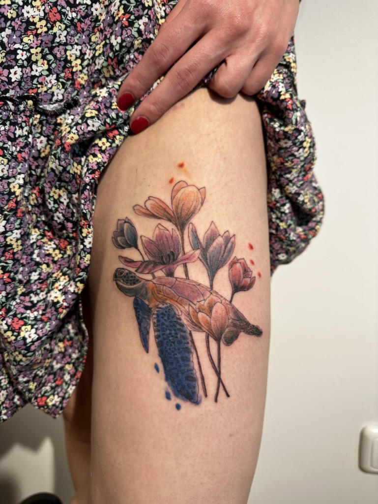 Tattoo of a sea turtle with blue flippers surrounded by pink flowers on a person's thigh.