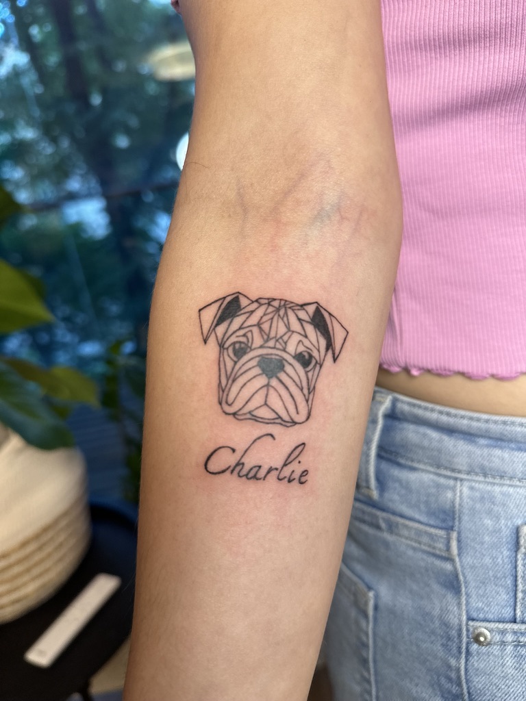 Geometric line art tattoo of a pug's face with the name Charlie below on a person's forearm.