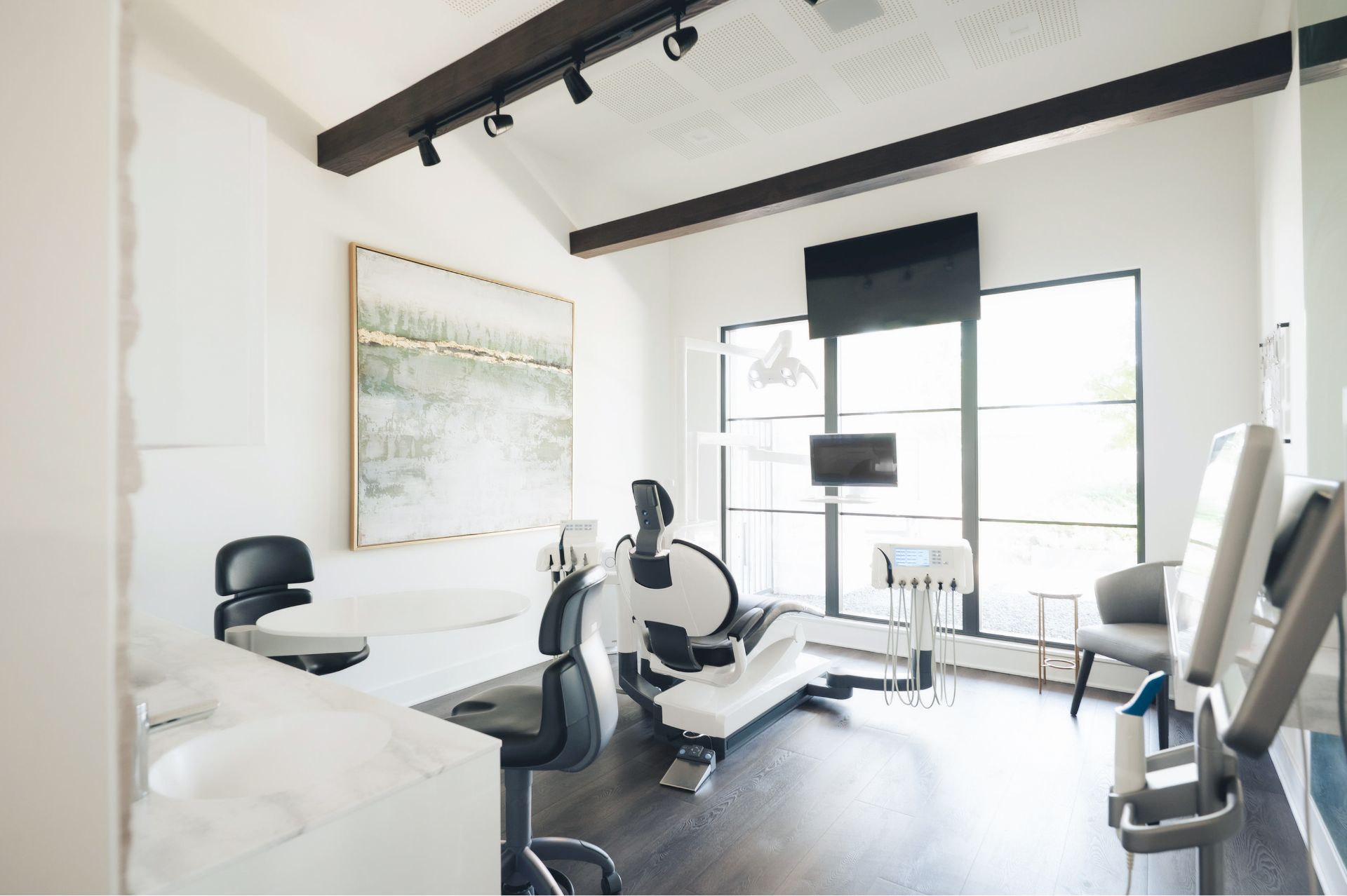Modern dental office with a dental chair, monitors, and black swivel stools near a large window.