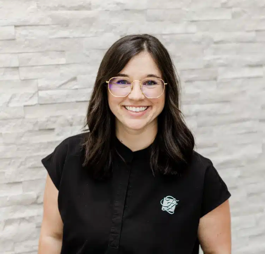 Smiling woman with medium-length dark hair wearing glasses and a black shirt with a logo, standing in front of a textured white stone wall.