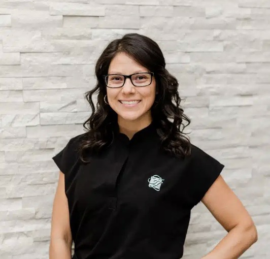 Smiling woman with glasses and wavy dark hair wearing a black sleeveless top, standing against a light gray textured wall.