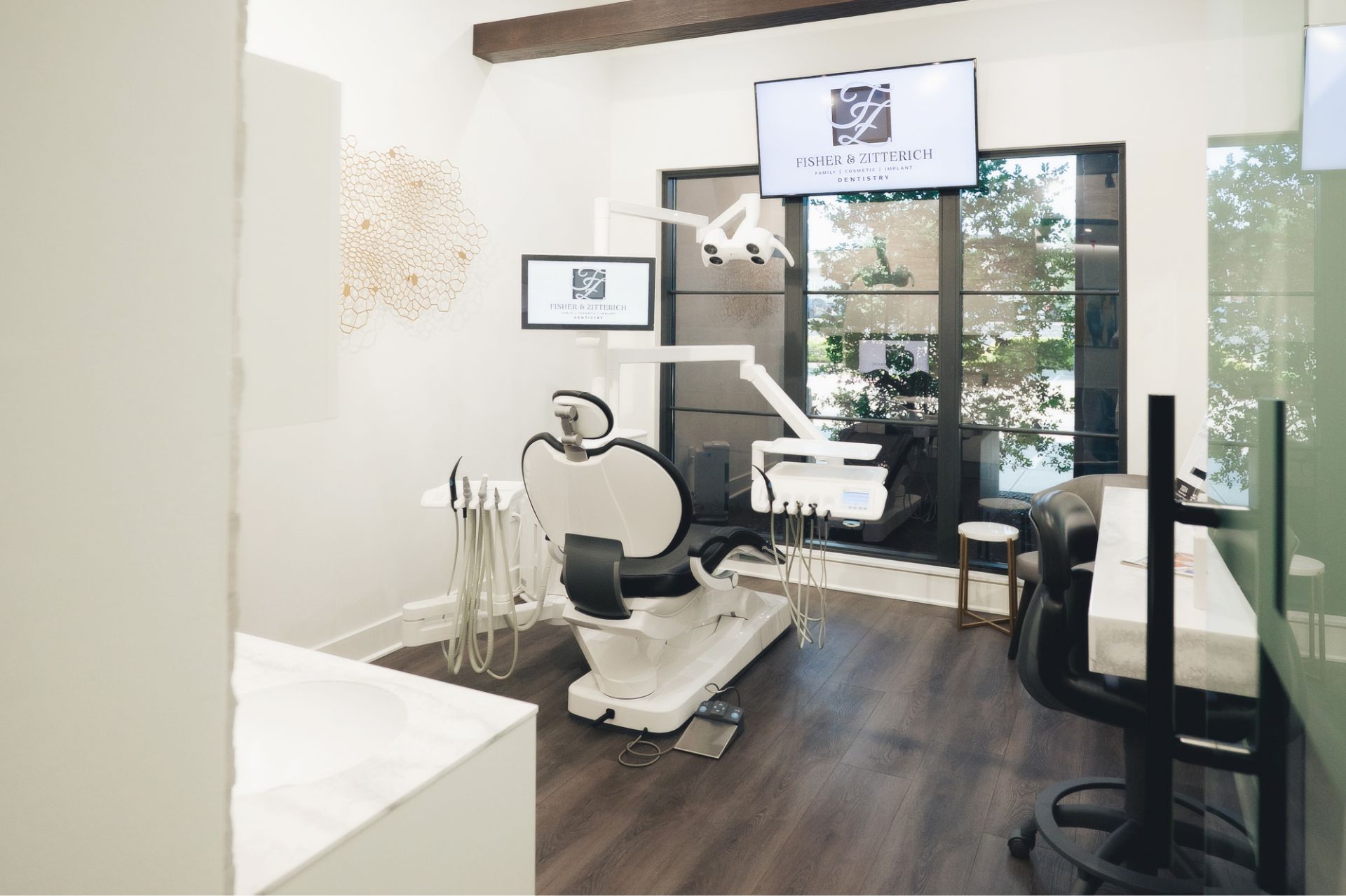 Modern dental examination room with a dental chair, equipment, two screens displaying Fisher & Zitterich Dentistry logo, and a large window showing outdoor greenery.
