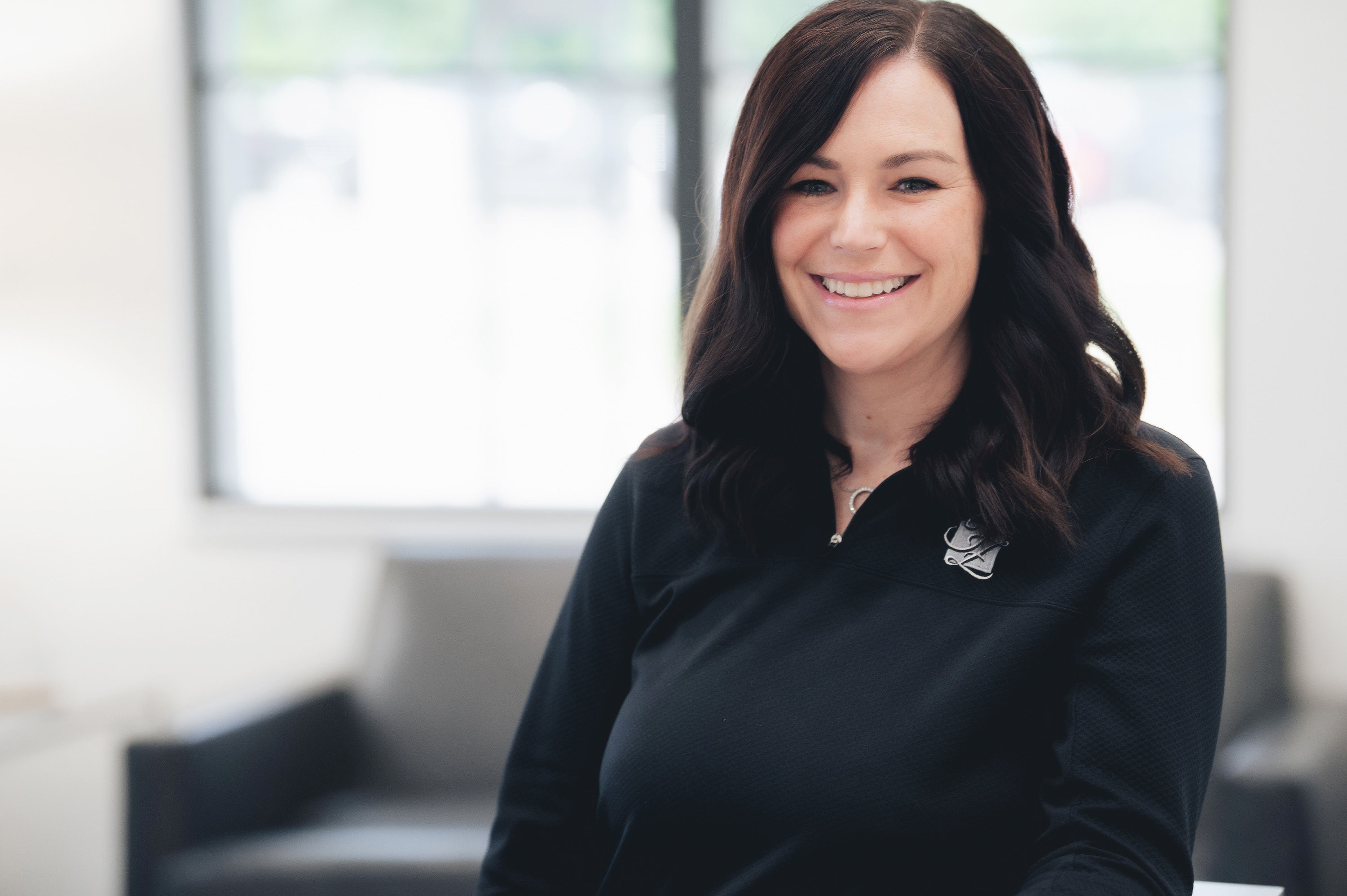 Smiling woman with dark hair wearing a black top with a logo, standing indoors near a window.