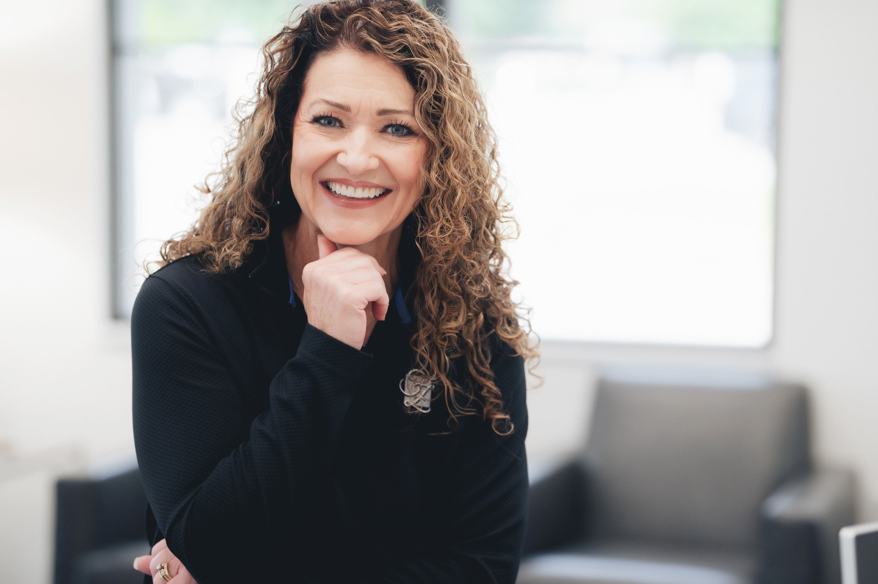 Smiling woman with curly hair wearing a black top sitting in a bright room with blurred furniture in the background.