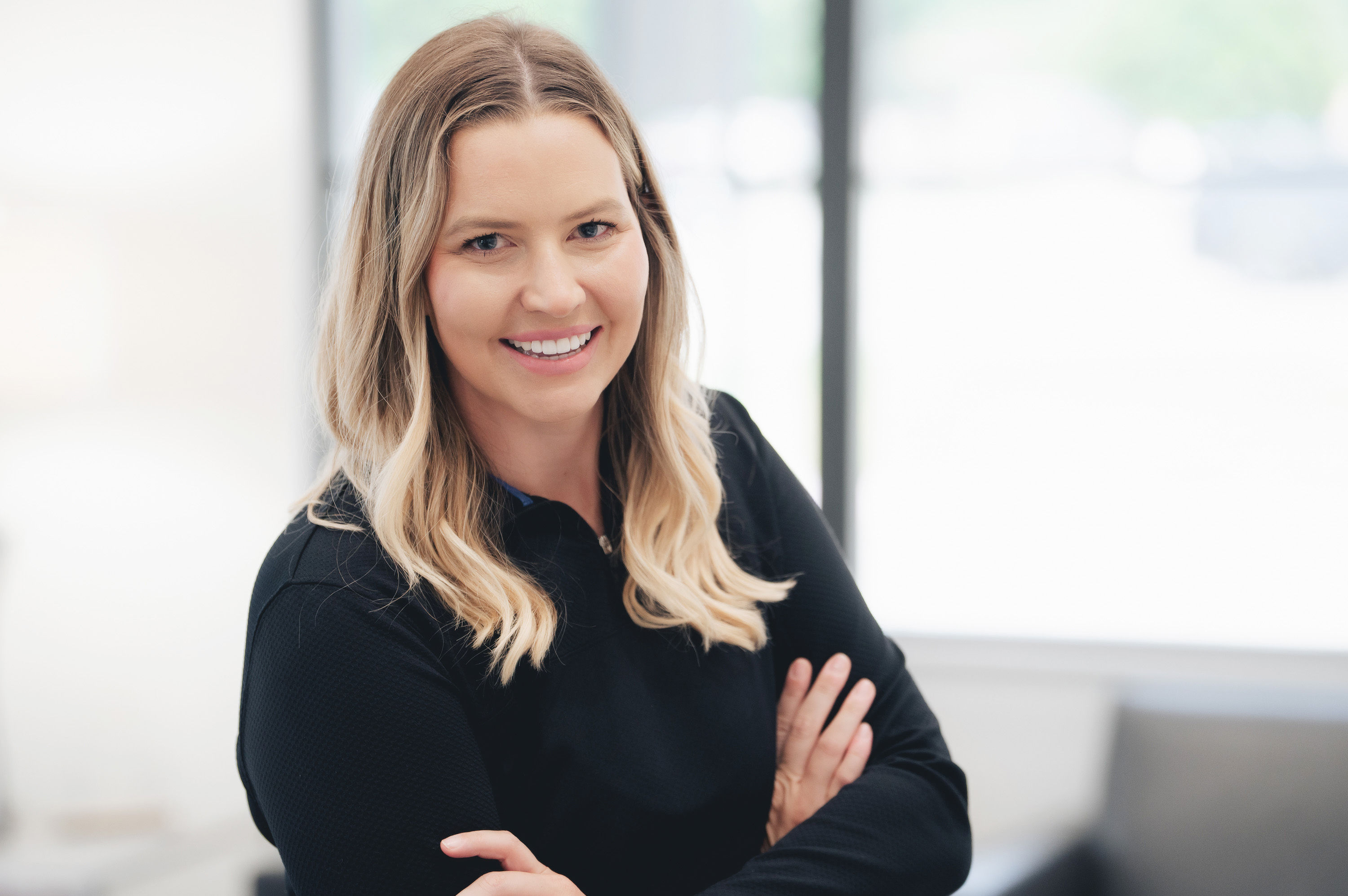 Smiling blonde woman with long hair and crossed arms wearing a black top in a bright room.