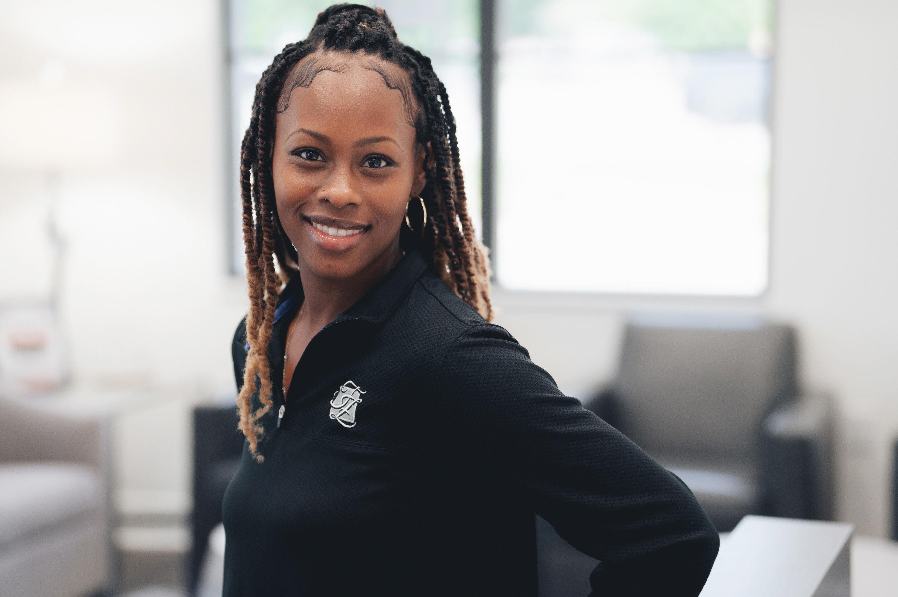 Smiling woman with braided hair wearing a black top standing in a bright, modern living room.