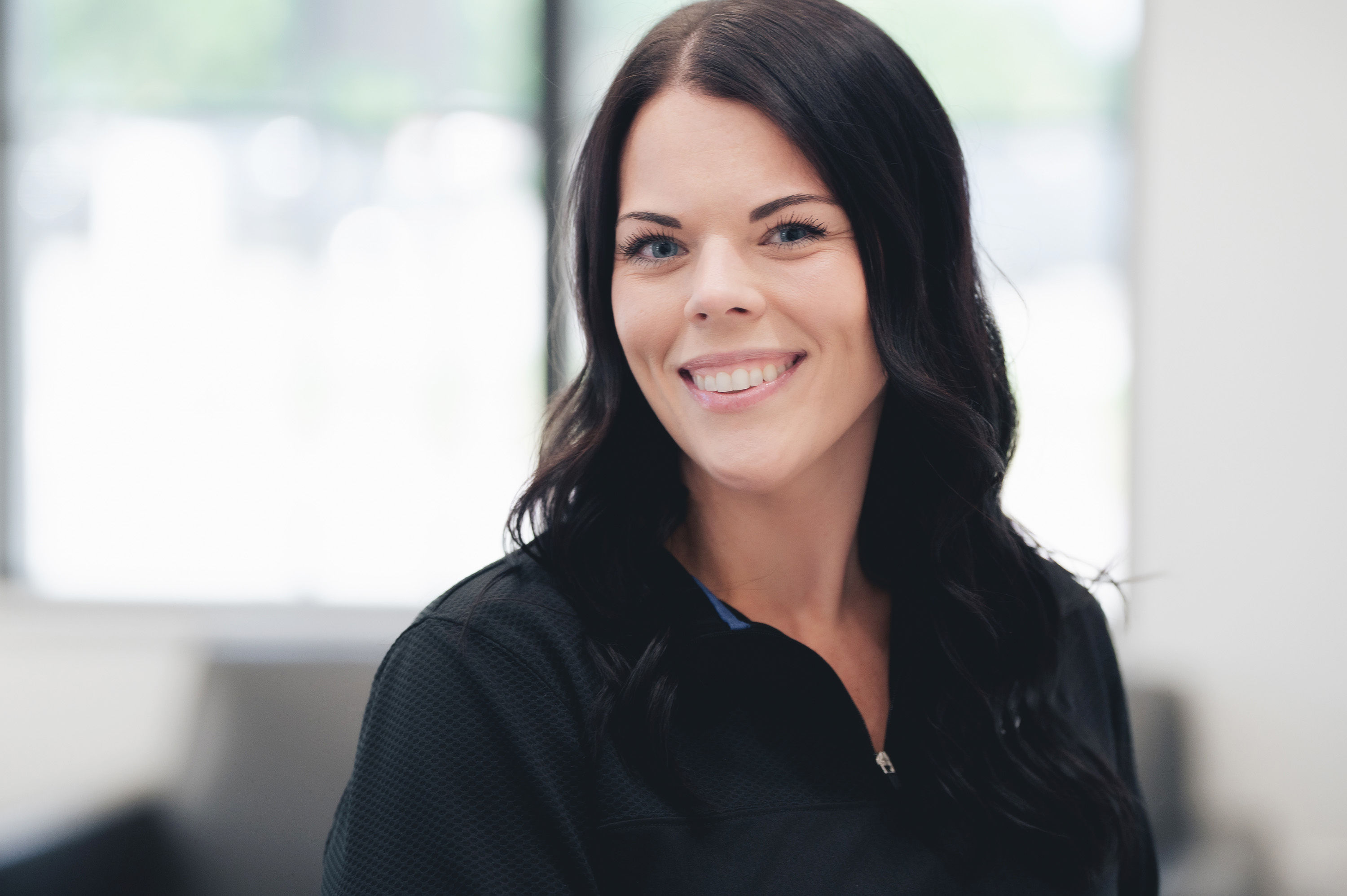Smiling woman with long dark hair wearing a black zip-up top in a bright indoor setting.