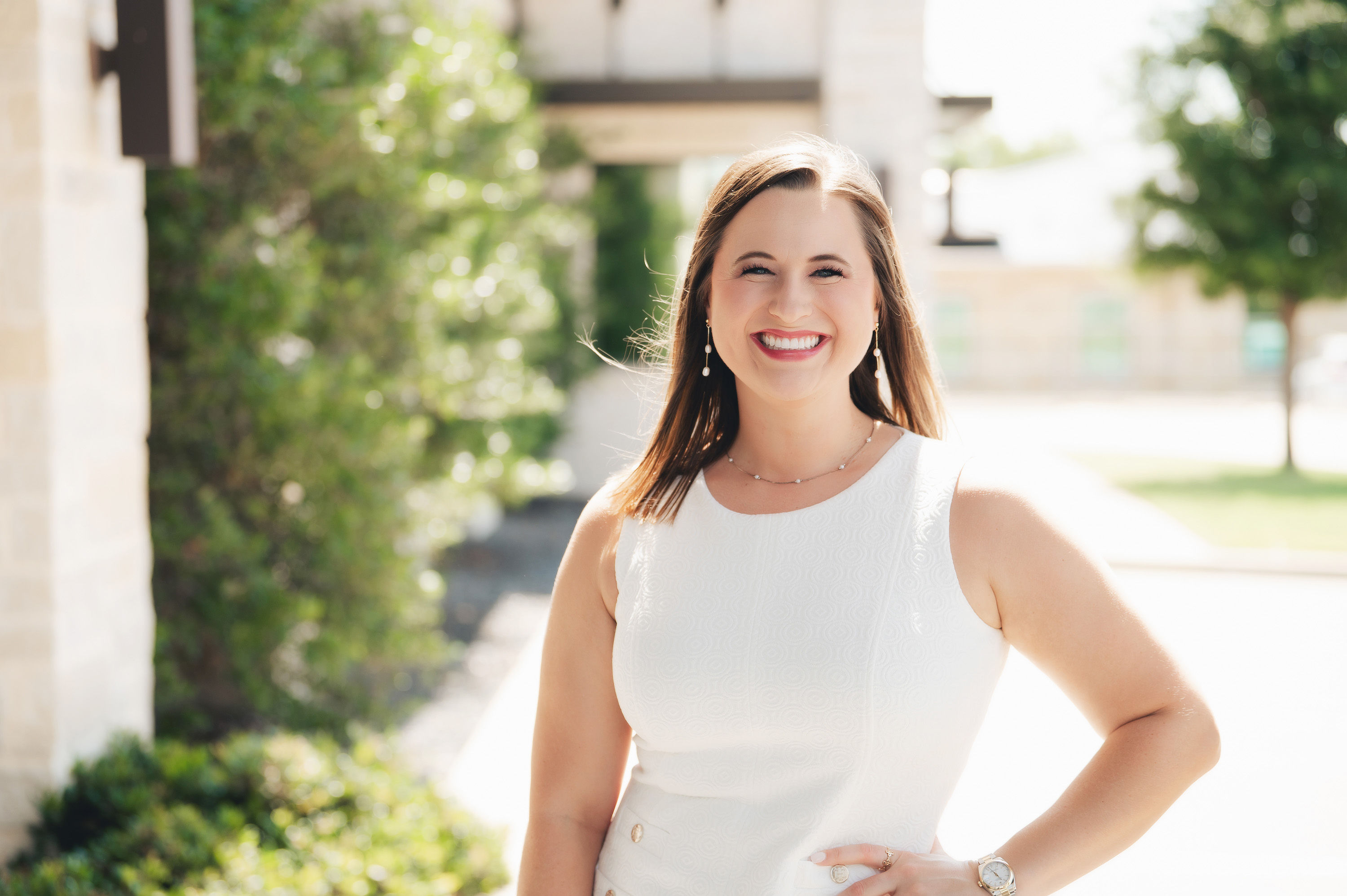 Smiling woman with long brown hair wearing a sleeveless white dress, standing outdoors with greenery and buildings in the background.