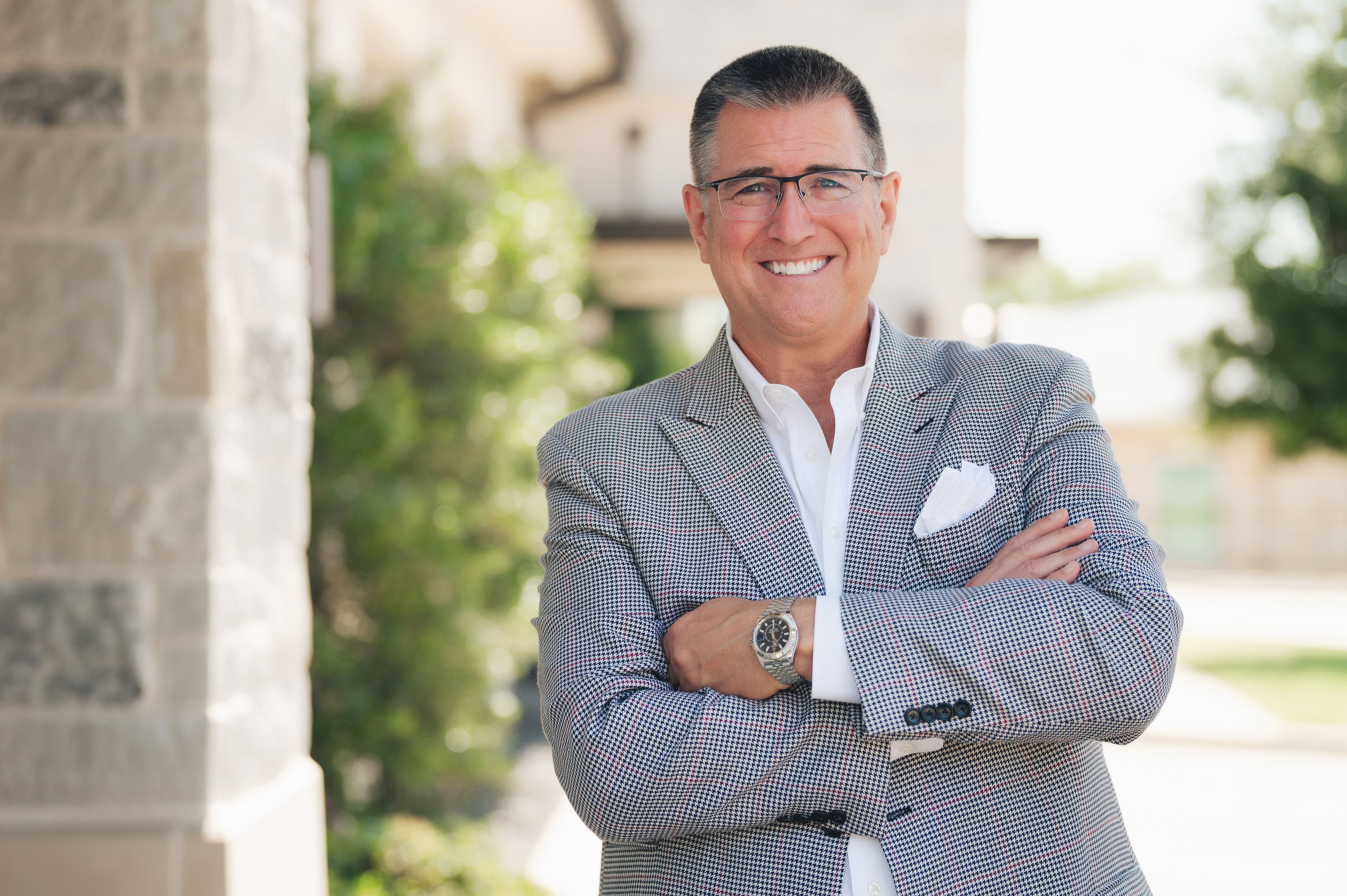 Smiling man wearing glasses and a checked blazer standing outdoors with arms crossed.