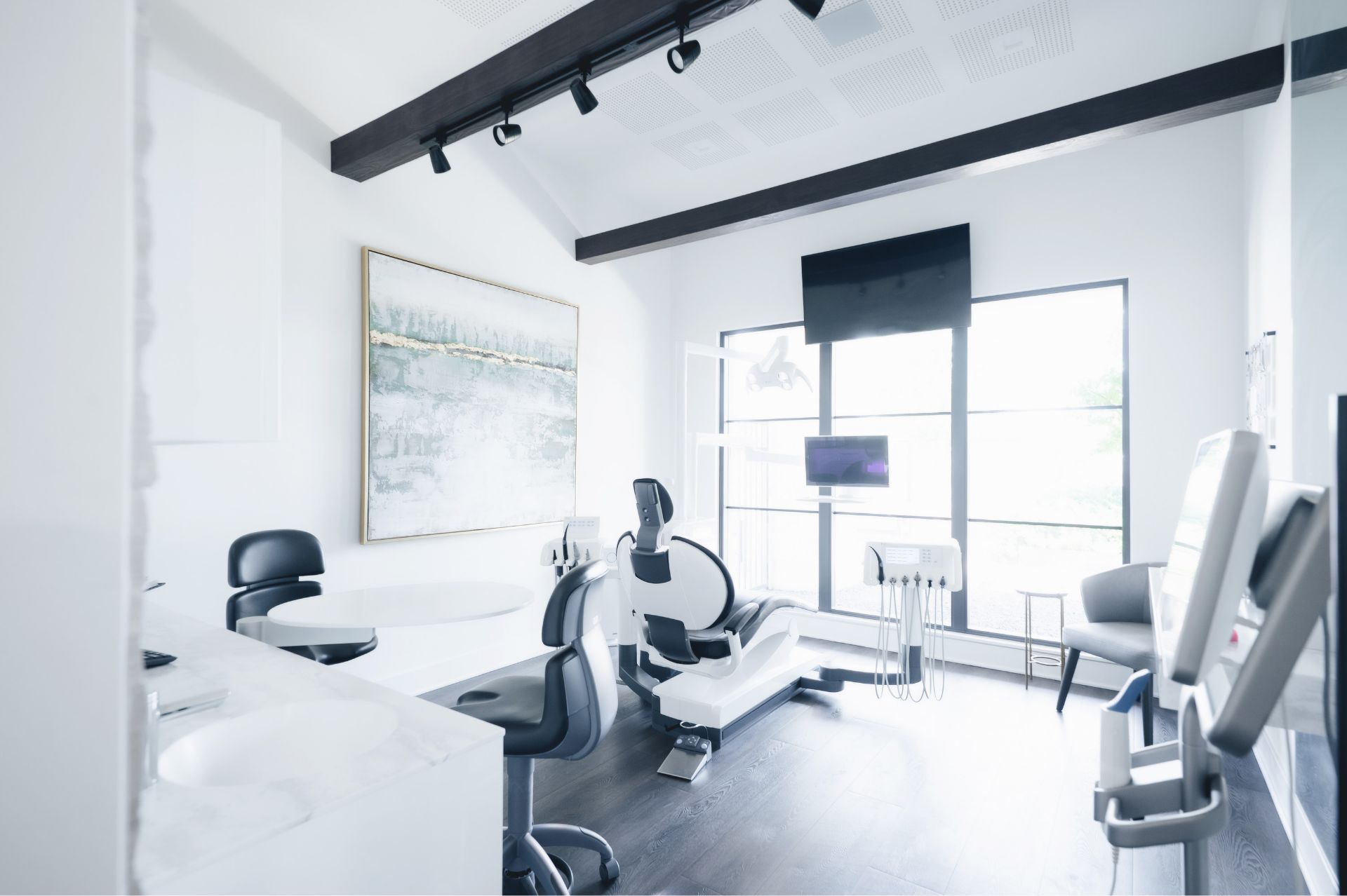 Bright modern dental examination room with dental chair, black stools, large window, and wall-mounted monitors.