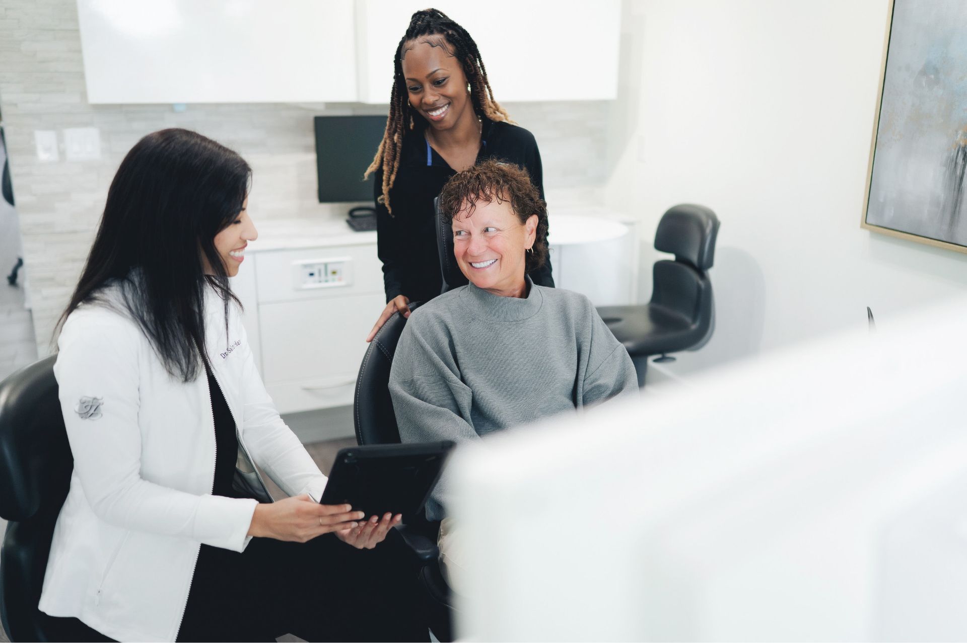 A healthcare professional shows information on a tablet to a seated woman while another woman stands behind them, all smiling in a clinical setting.