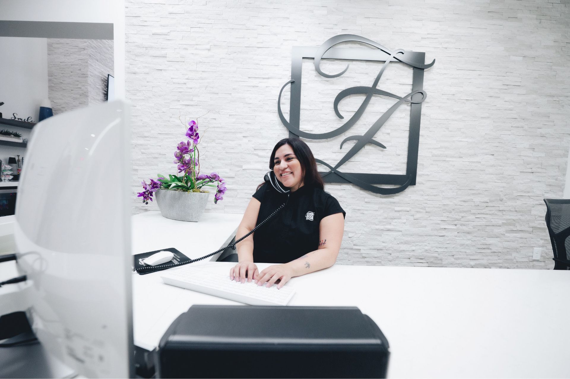 Smiling receptionist talking on a corded phone and typing at a white desk with a purple orchid plant and a logo on the wall behind her.