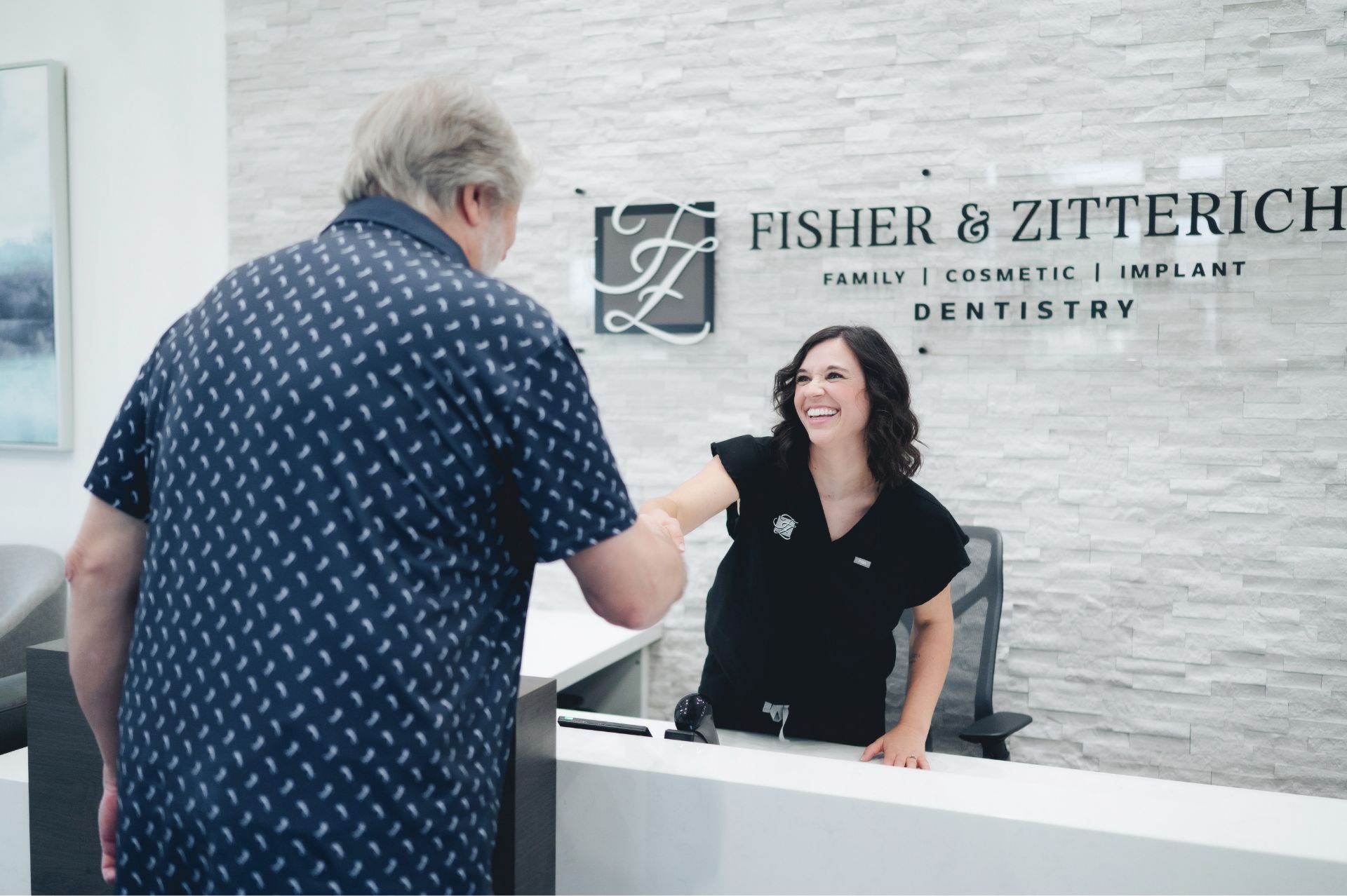Smiling dental receptionist shaking hands with a male patient at the front desk of Fisher & Zitterich Dentistry.