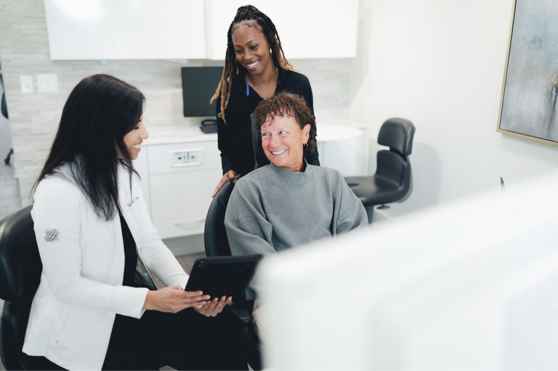 Smiling female healthcare professional showing a tablet to a seated middle-aged woman while a young woman stands behind them in a bright medical office.