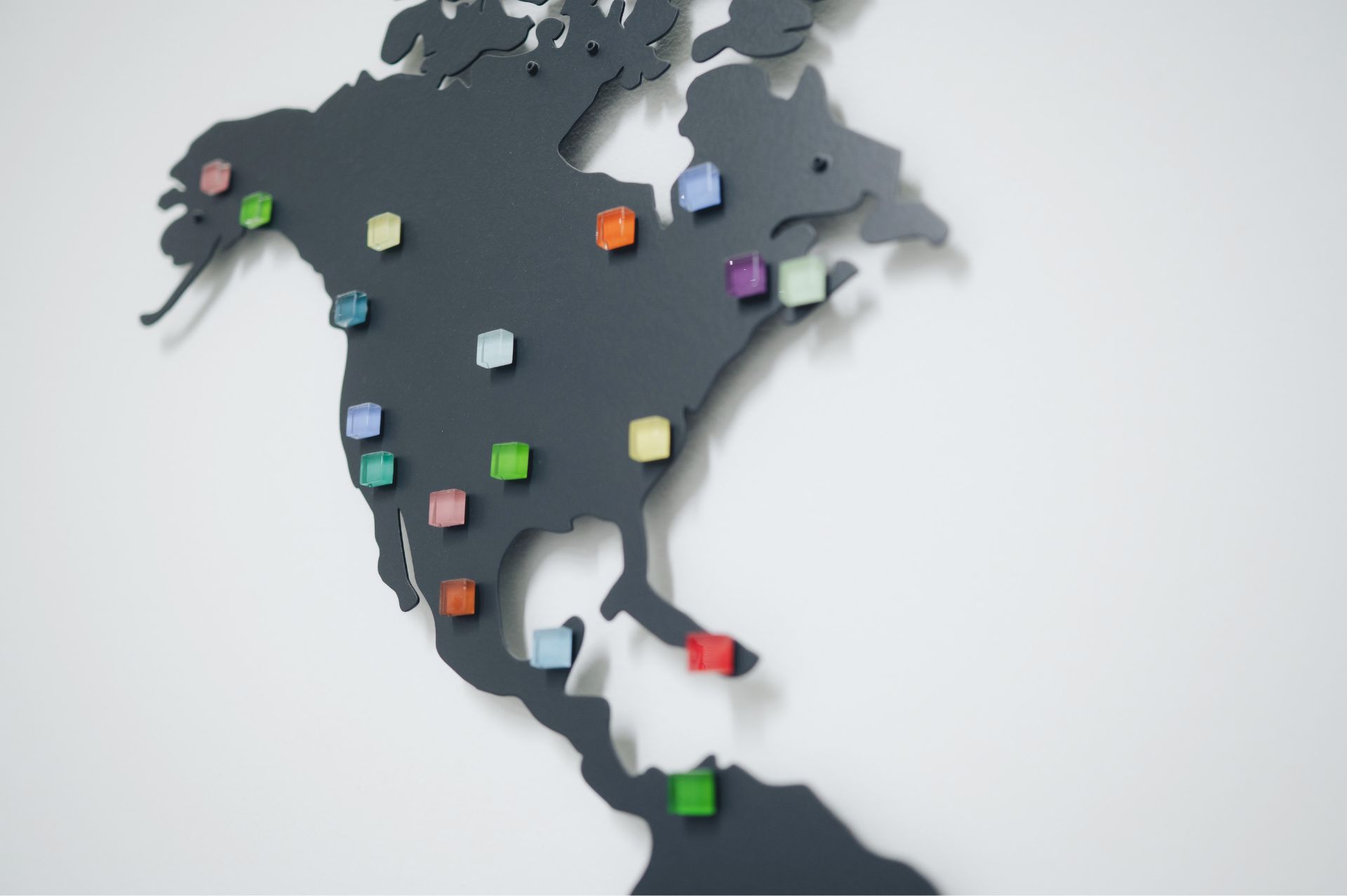 Black stylized map of North America mounted on a white wall with colorful square pins marking various locations.