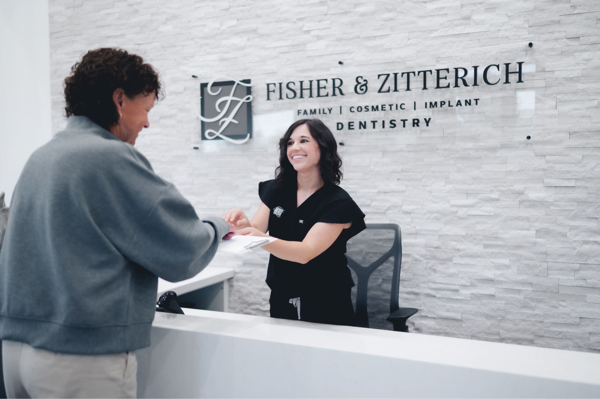Smiling dental receptionist handing a clipboard to a patient at Fisher & Zitterich Dentistry reception desk.