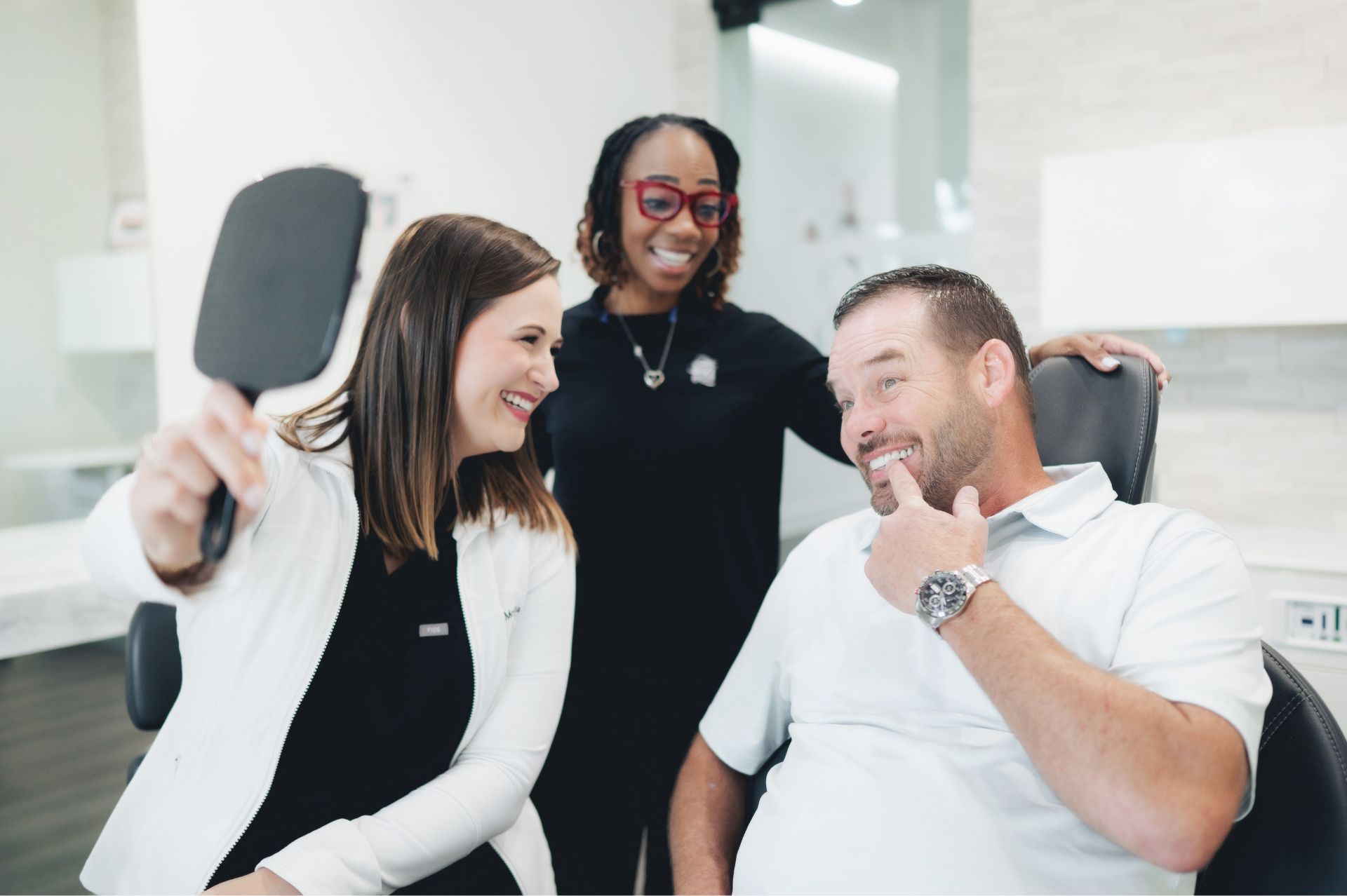 Smiling woman in a white coat holding a mirror while a man in a dental chair points at his teeth, with another woman standing behind them smiling.